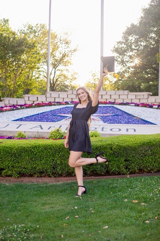 Graduate celebrates by landscaped university entrance with decorative flowers and flags.