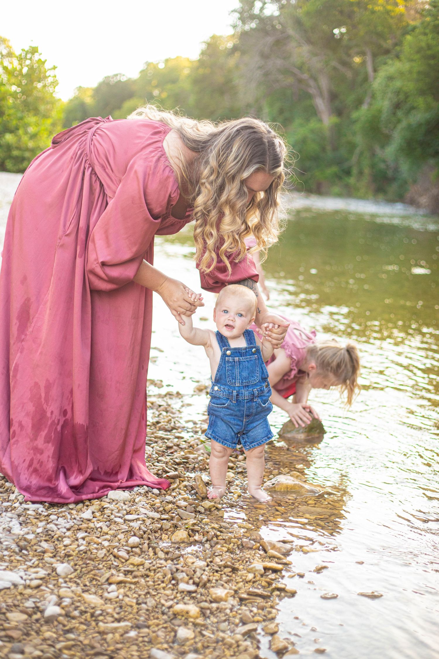Parent and children splash and play in shallow river water while wearing pink and denim outfits.