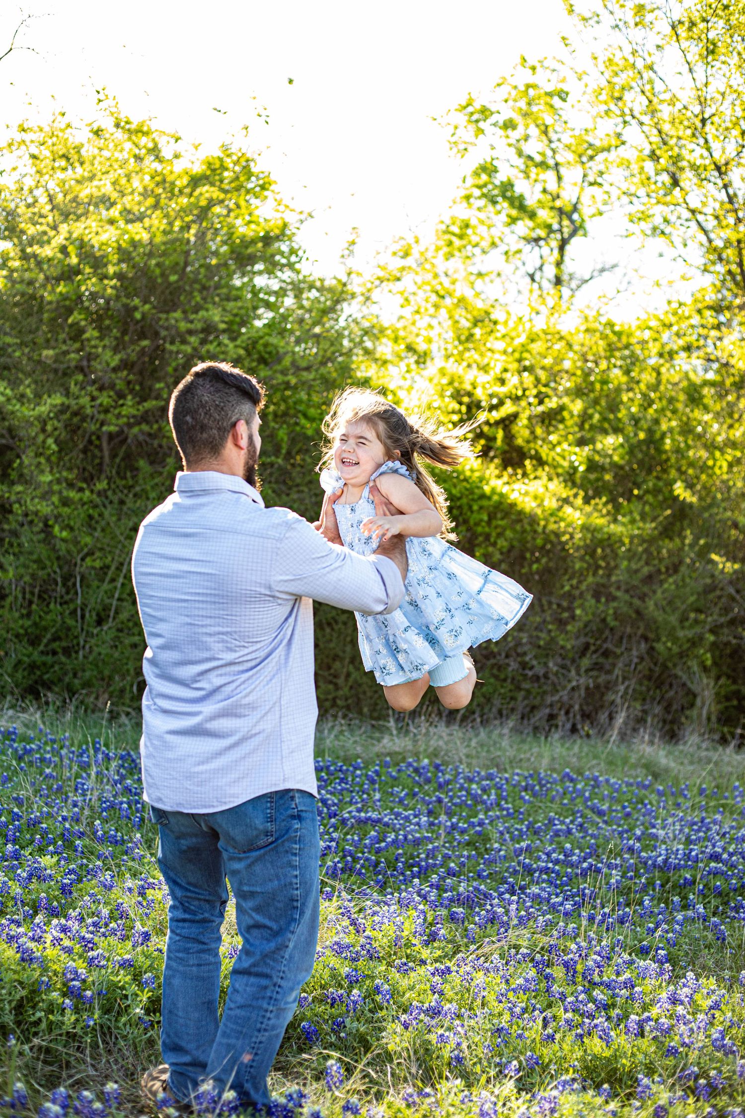 A father joyfully lifts his smiling daughter in a field of bluebonnets with lush green trees in the background.
