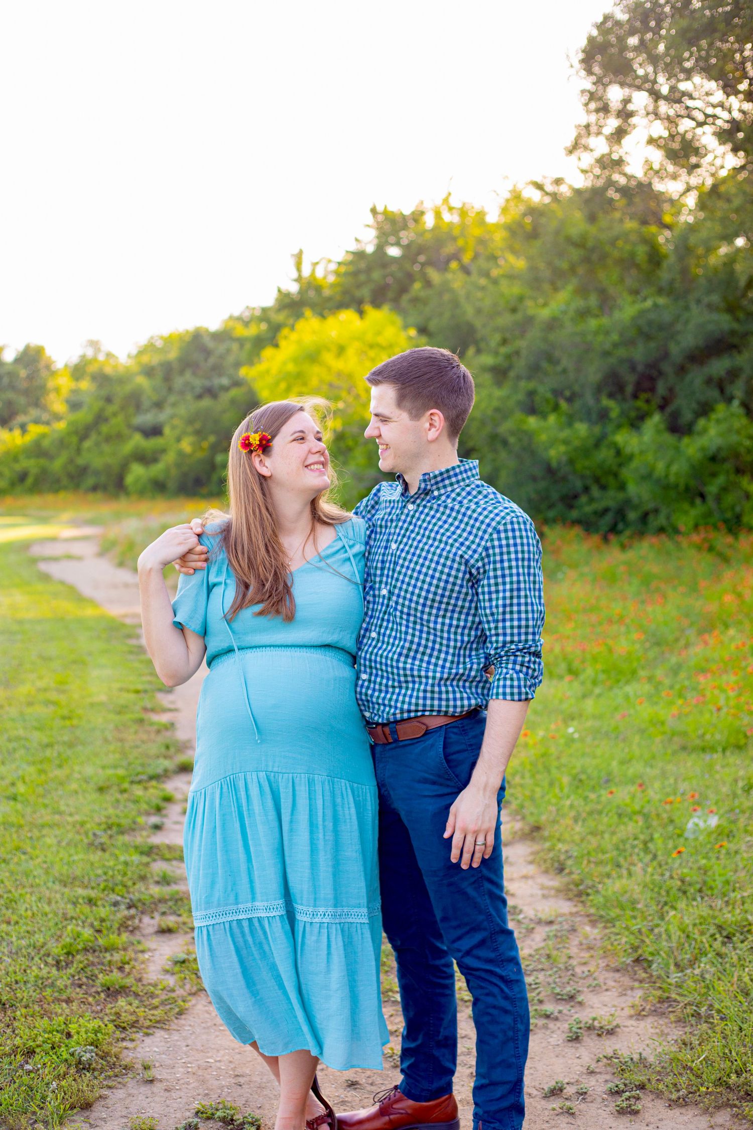 Pregnant woman in blue dress and man in plaid shirt share a loving glance on a sunny outdoor path.