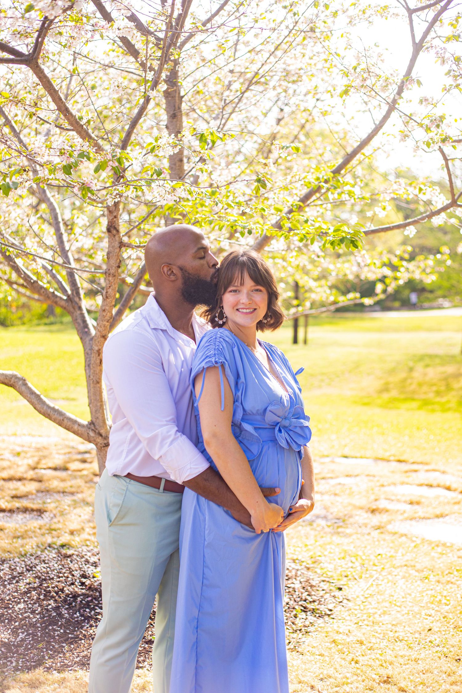 Couple embracing under blooming trees in a sunlit garden setting wearing blue dress and casual attire.