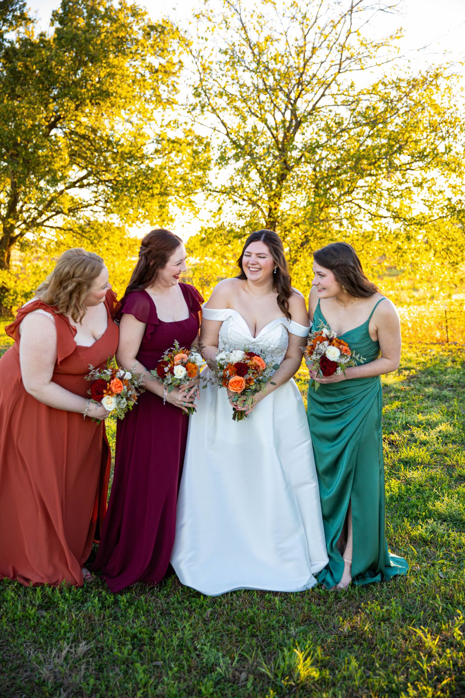 Bride in white gown with three bridesmaids in rust, burgundy, and green dresses holding bouquets outdoors.
