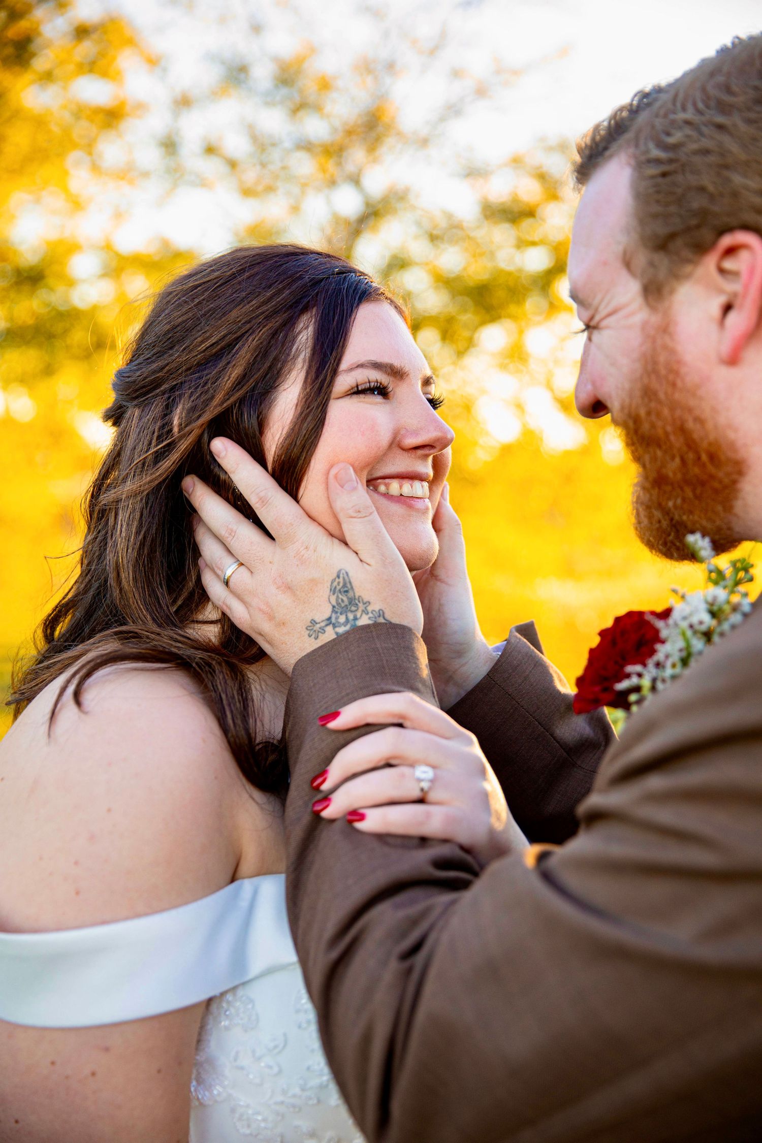 A smiling bride with red nails and a diamond ring laughs as her red-bearded groom tenderly cups her face outdoors in fall.
