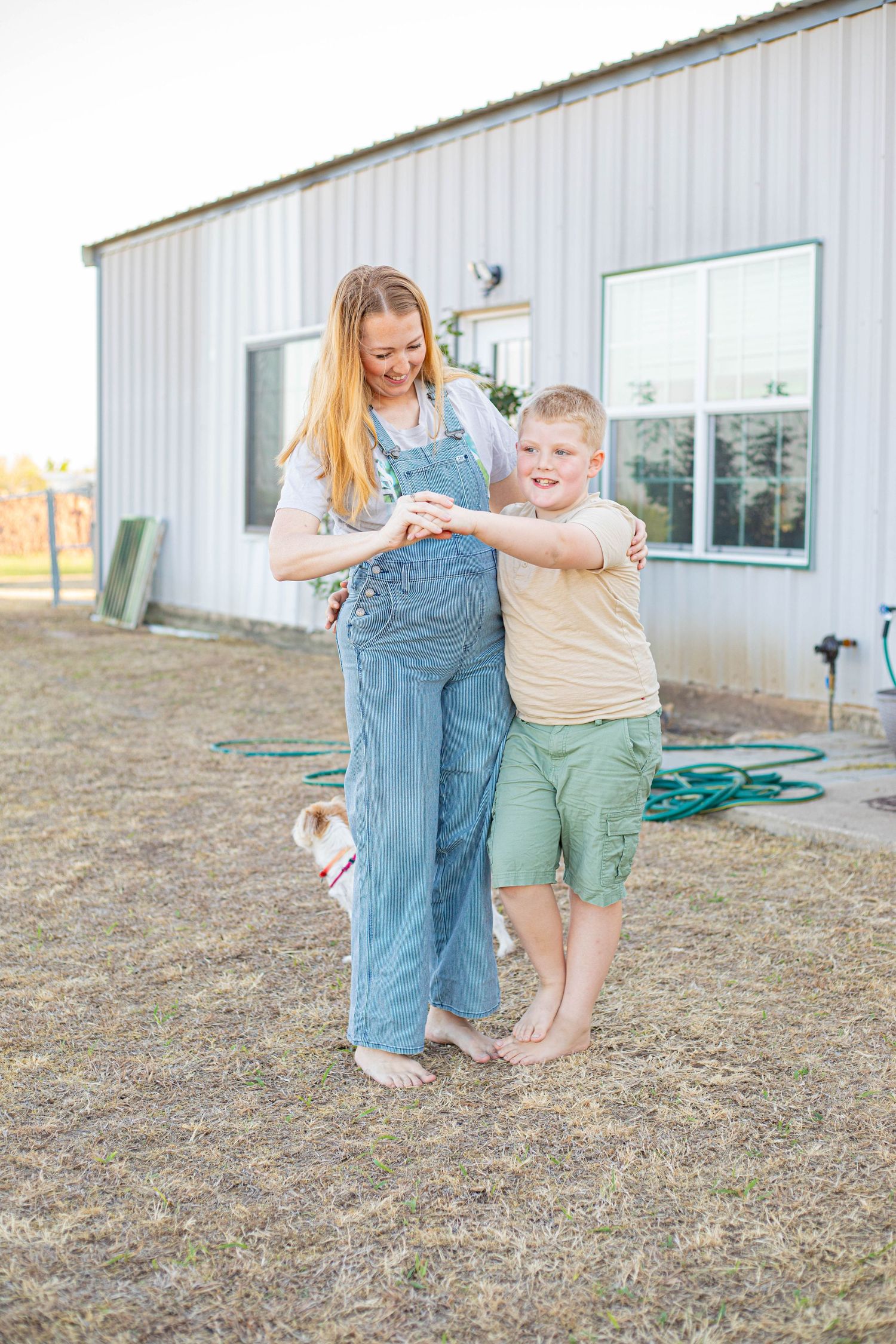 Pregnant woman in denim overalls smiling with young boy hugging her belly outdoors near a white barn.