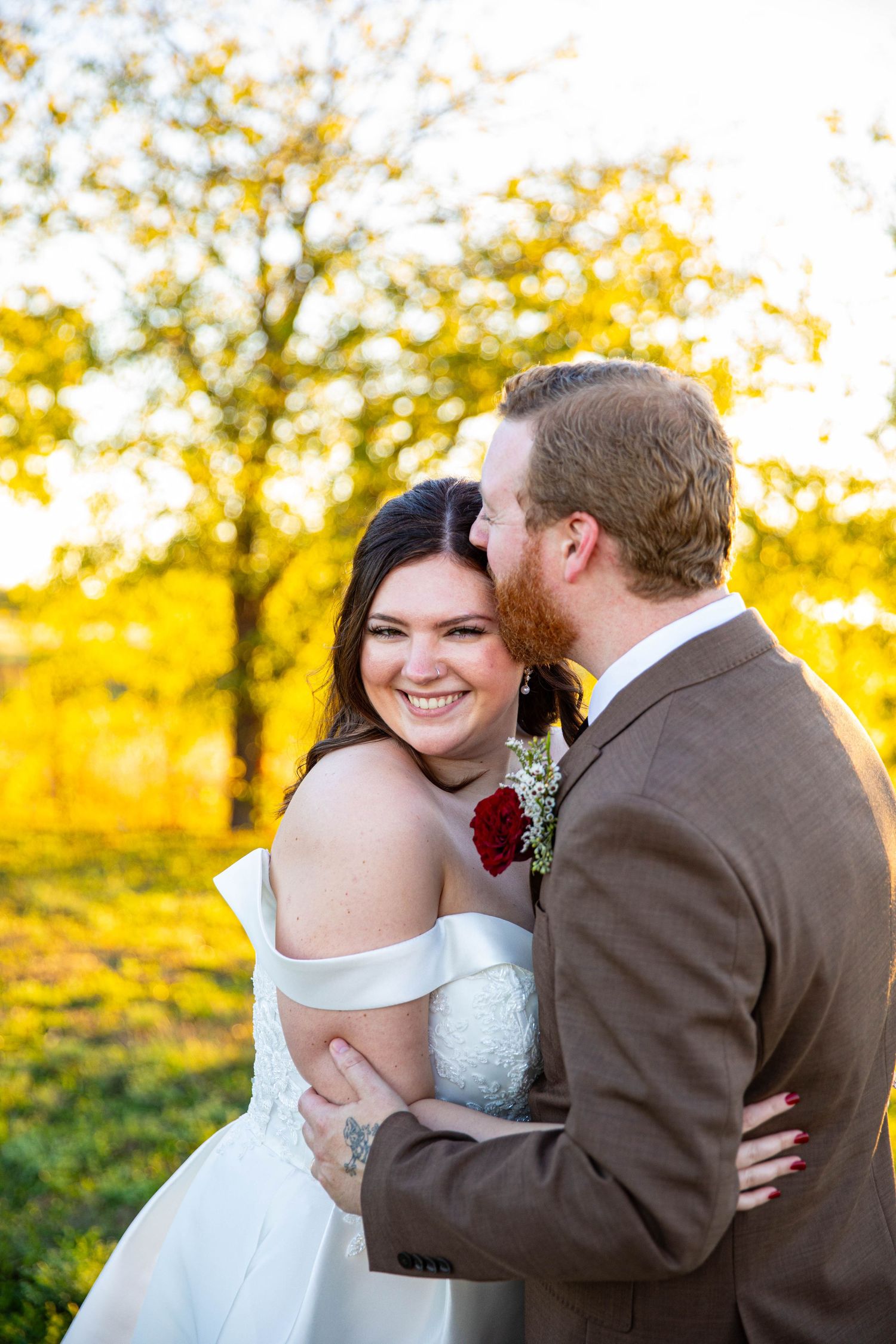 Bride in off-shoulder gown smiles as groom in brown suit whispers in her ear outdoors during golden hour.