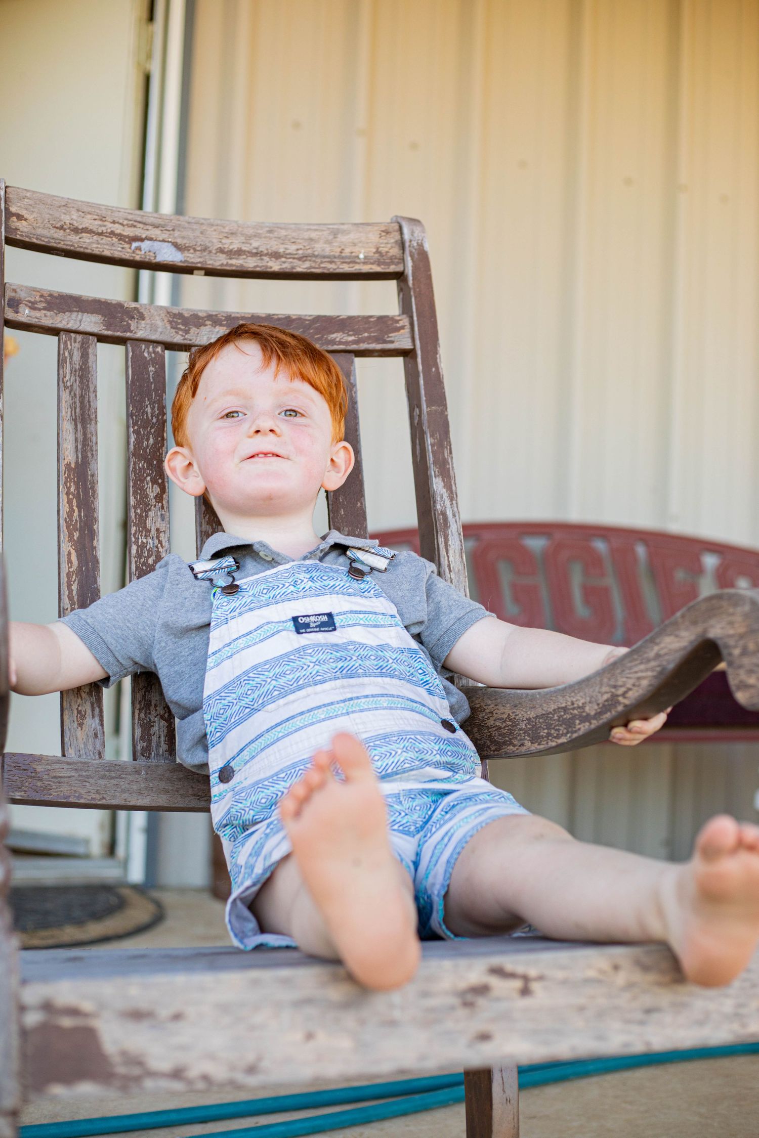 Red-haired toddler boy sitting relaxed in a rustic wooden chair on a porch, wearing striped overalls.
