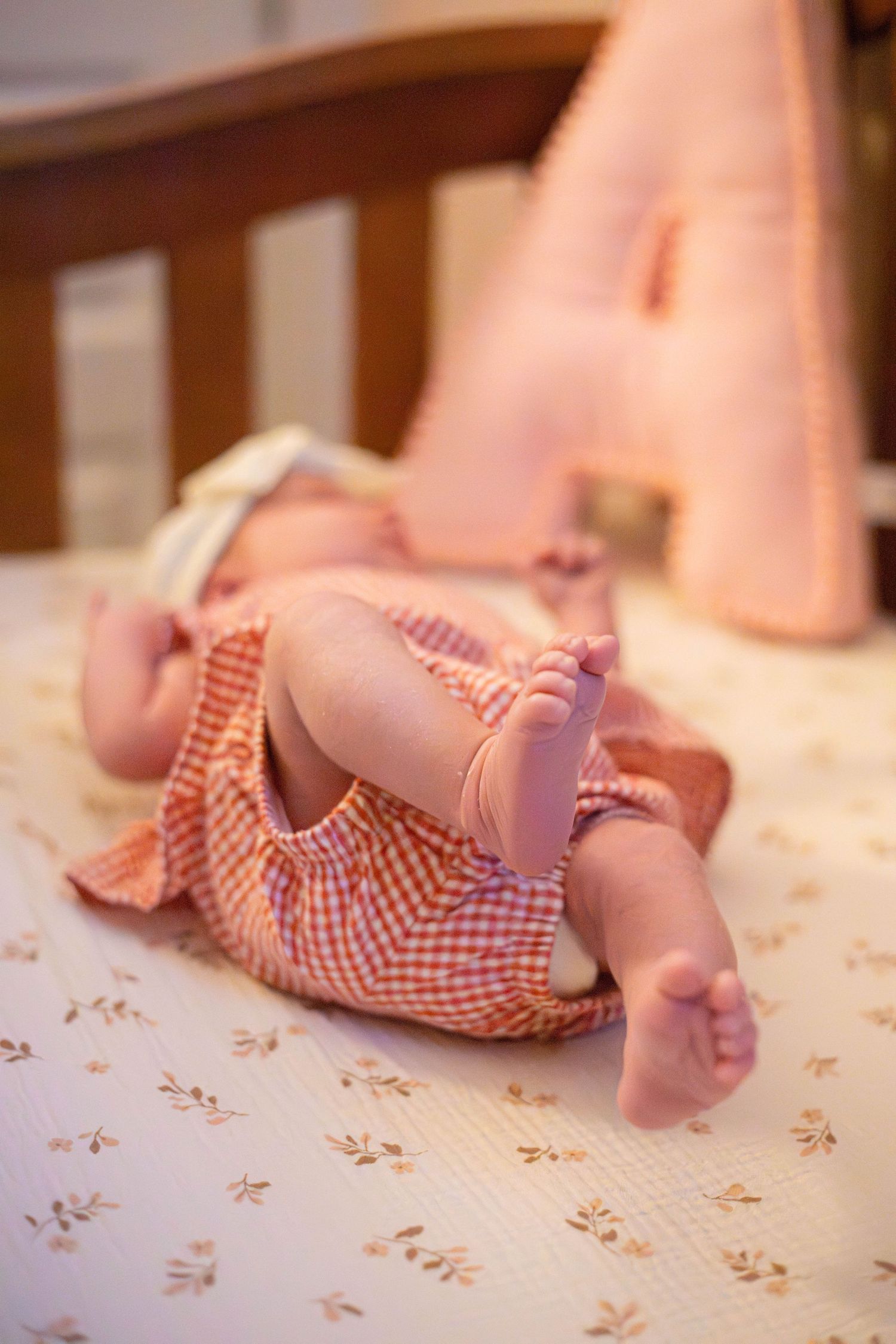 Newborn baby lying on back in red gingham outfit, tiny feet raised, resting in a wooden crib with pink letter A pillow.