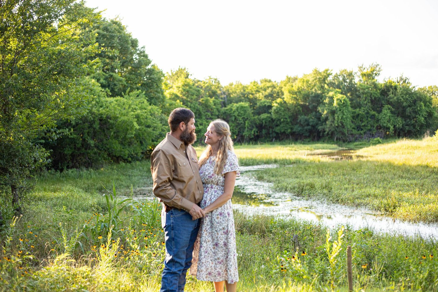 Couple embracing in a lush green meadow near a pond during golden hour in a romantic outdoor setting.