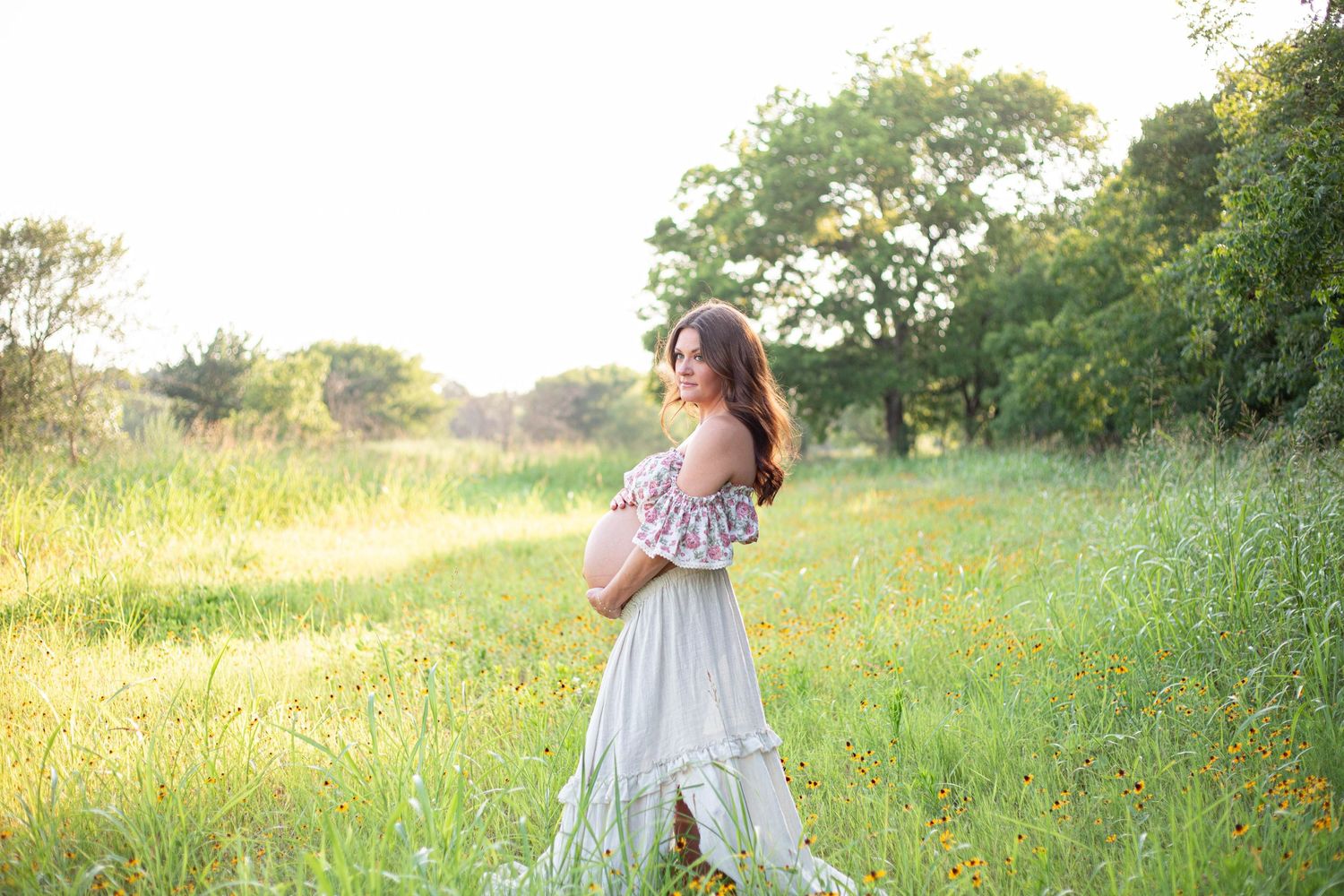 Pregnant woman in white floral dress cradling baby bump in sunlit green meadow during golden hour maternity photoshoot.