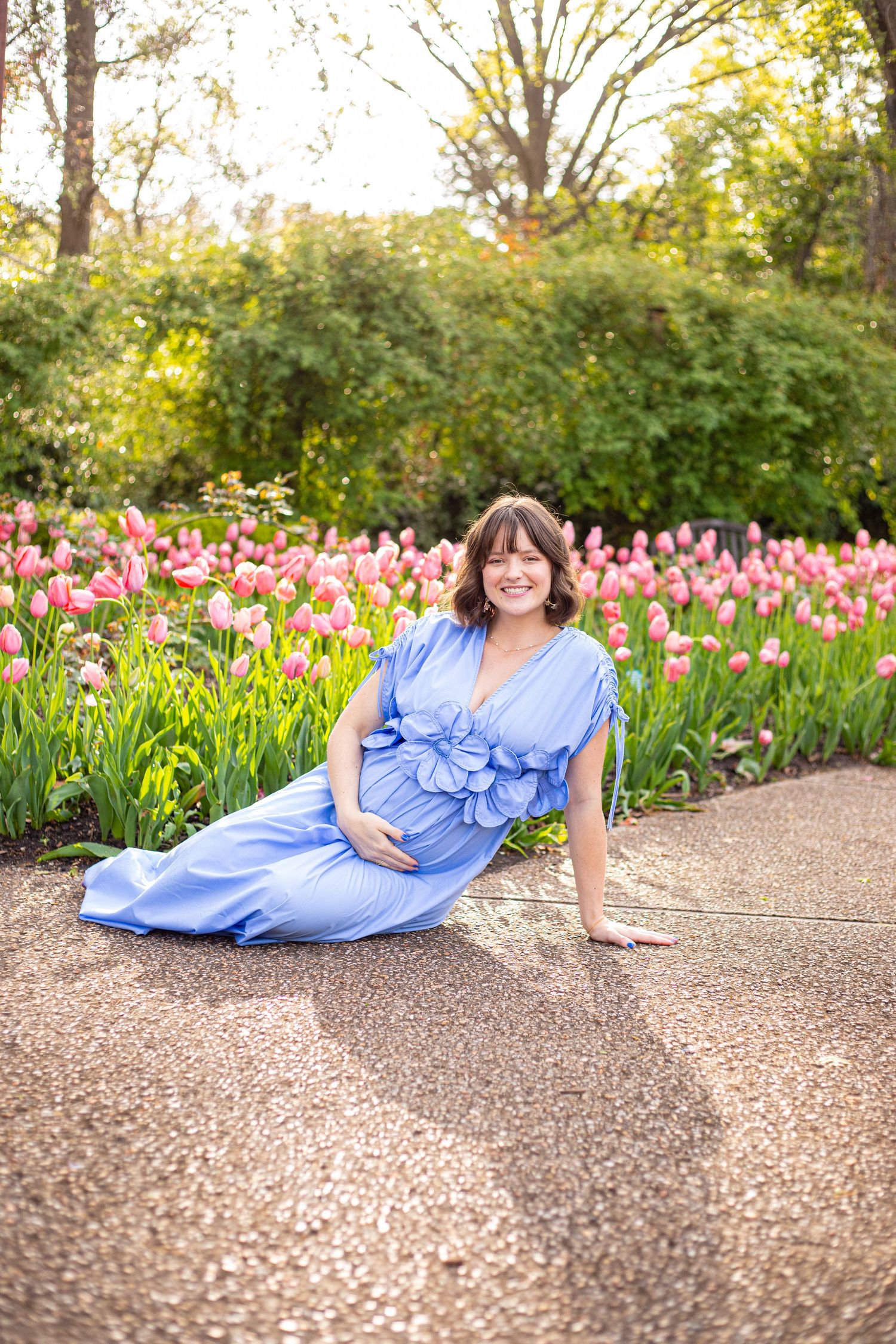 Woman in blue dress reclines on garden path surrounded by pink tulips and greenery.