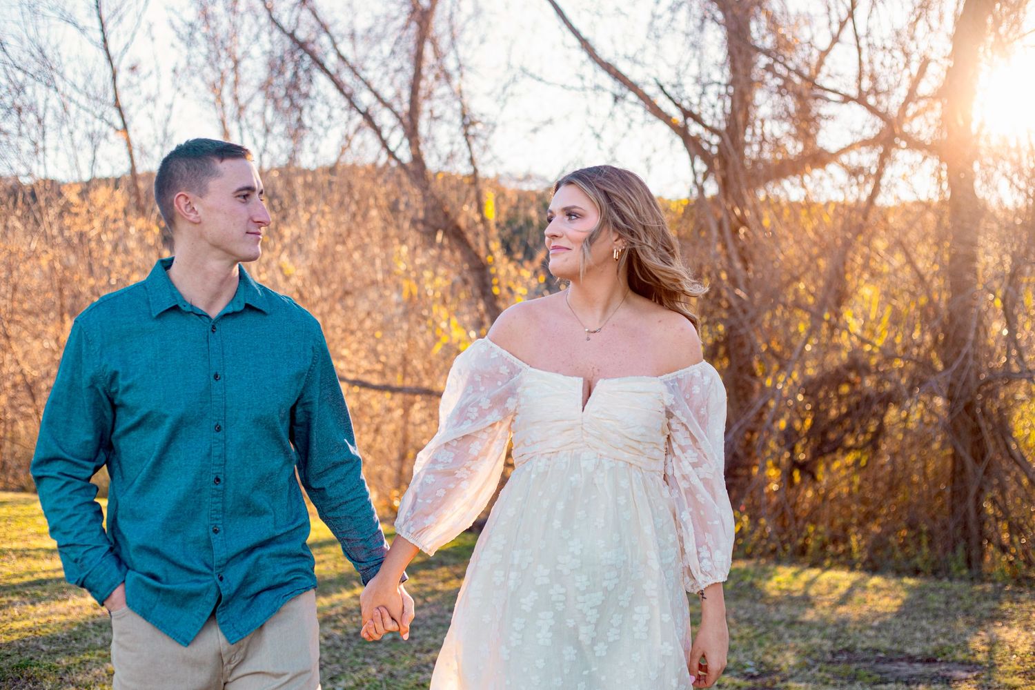 A couple walks hand in hand during sunset with fall foliage in the background.