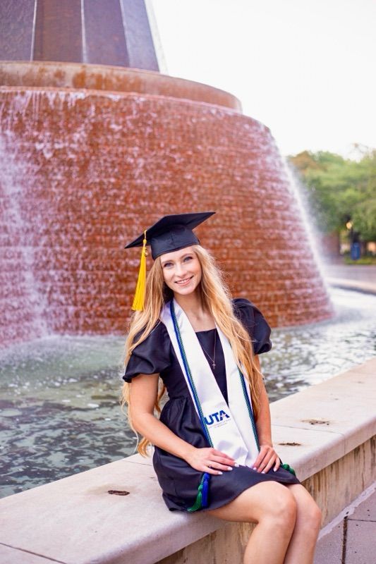 Graduate in academic regalia sits by a circular brick fountain on campus.