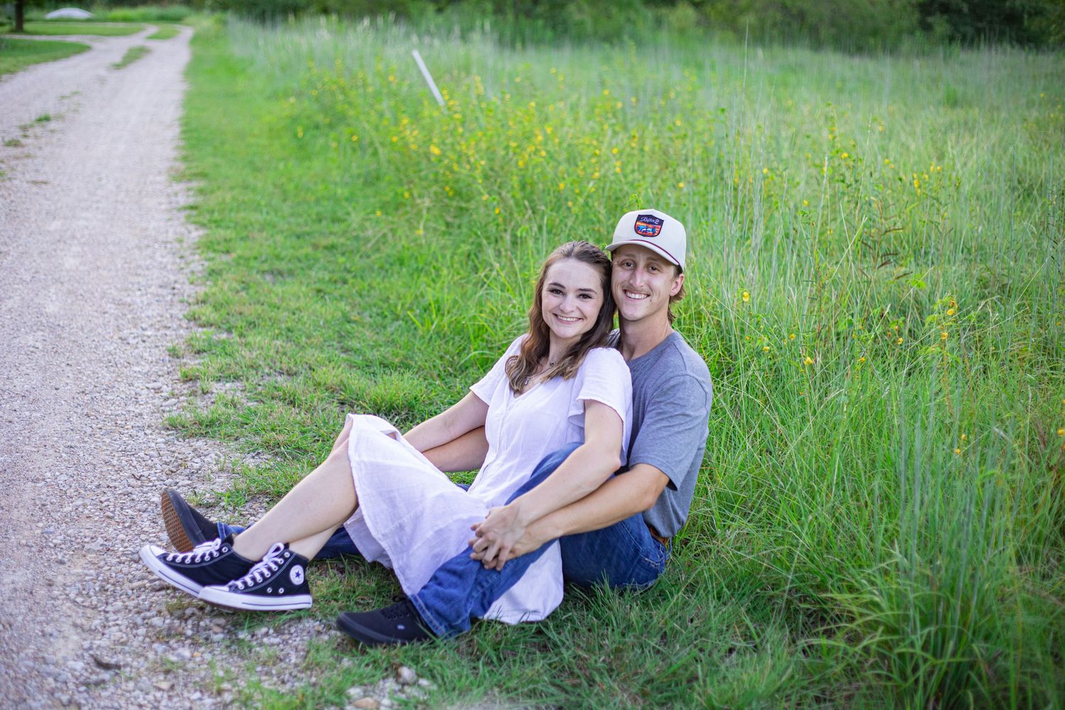 A couple sits together on a rural path surrounded by green grass and wildflowers.