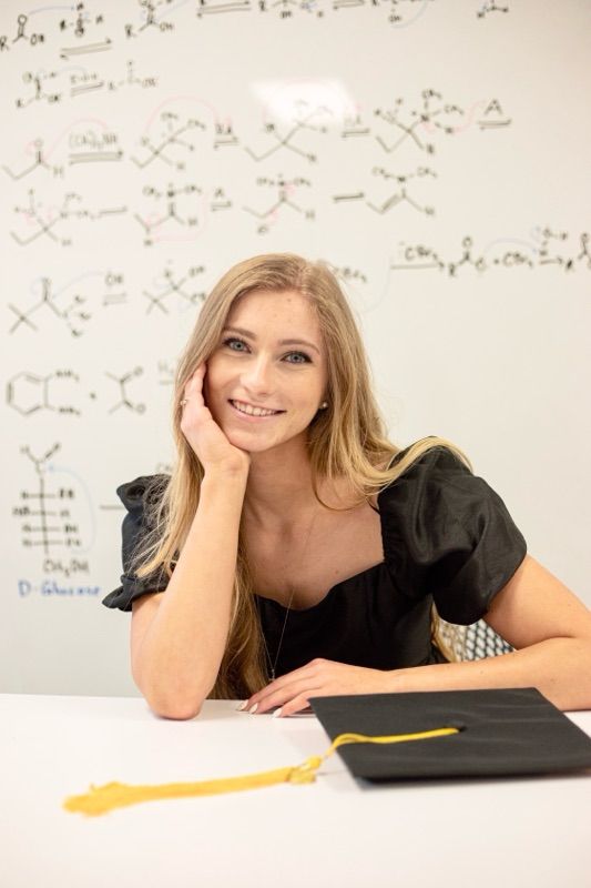 Academic setting with complex molecular diagrams and formulas written on whiteboards in the background.