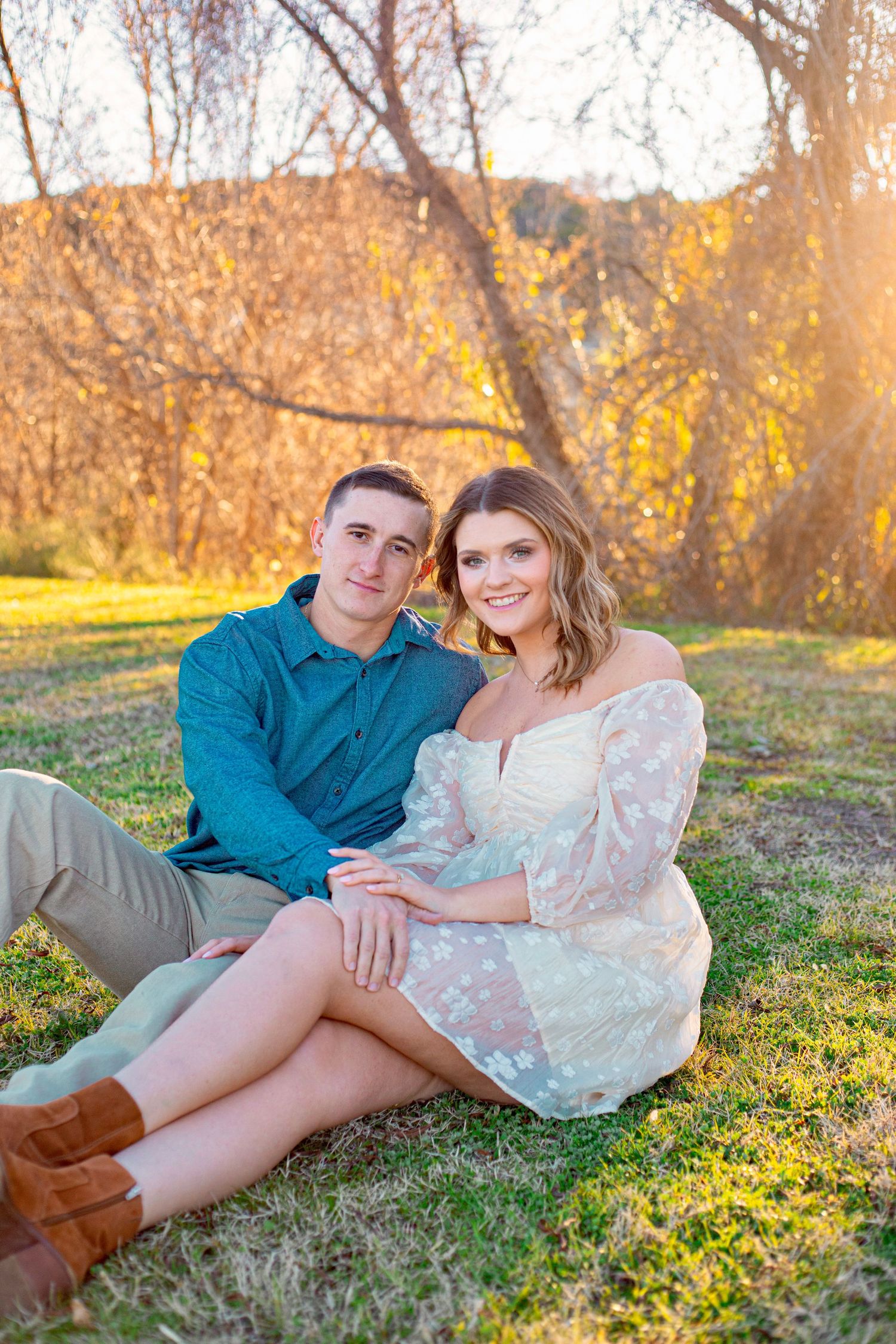 A couple sits together on grass during golden hour with autumn trees in the background.