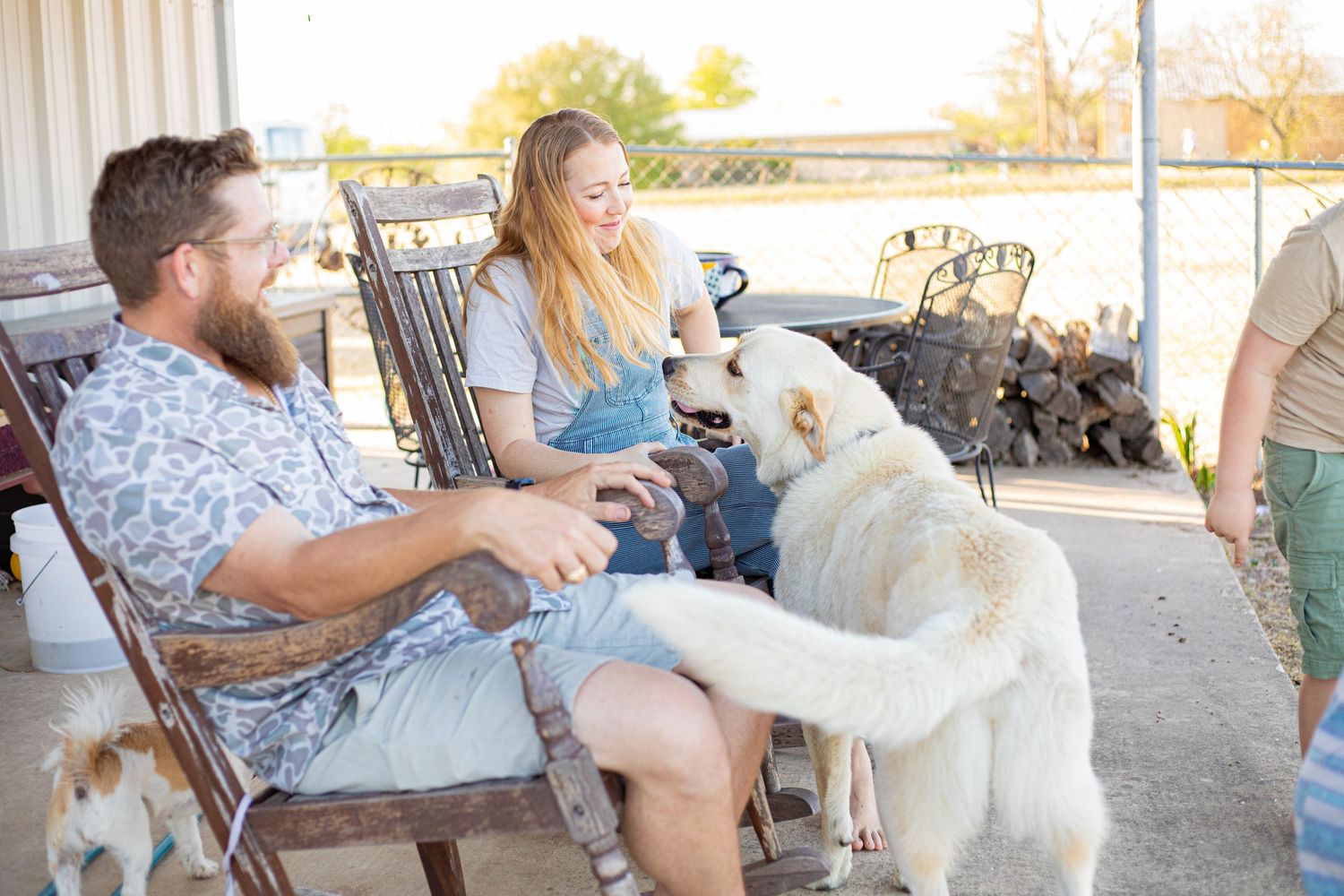 Couple laughing in rocking chairs on a farm porch while a large white dog playfully interacts with them.