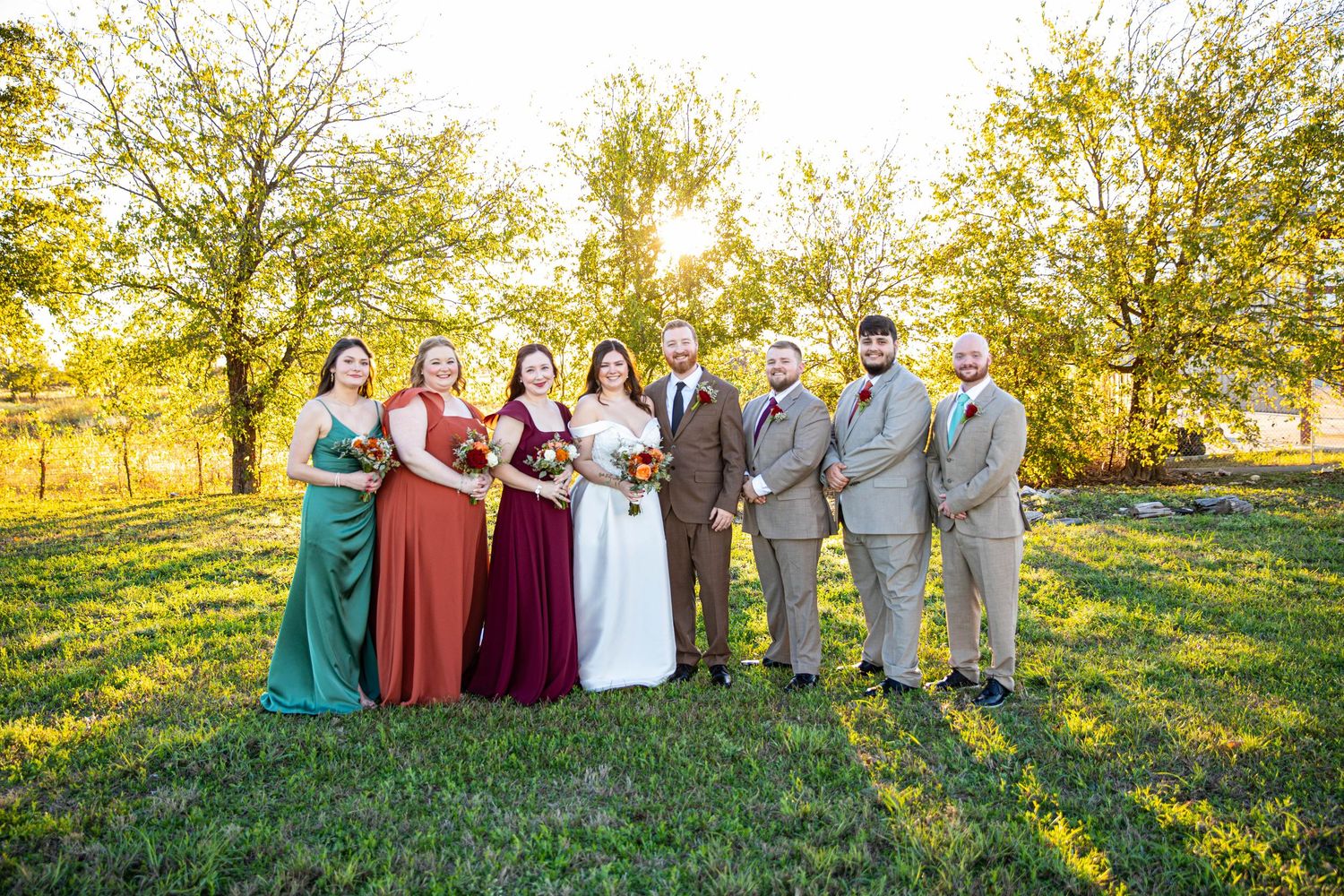 Wedding party posing outdoors in golden hour light, bridesmaids in green and red dresses, groomsmen in gray suits.