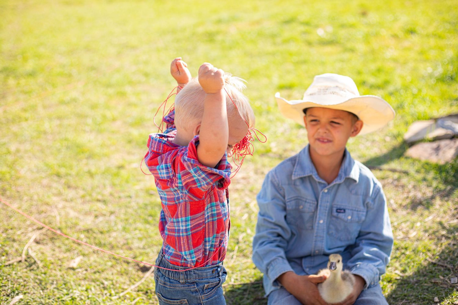 A child in plaid and an adult in denim sit together outdoors in bright sunlight on grassy ground.