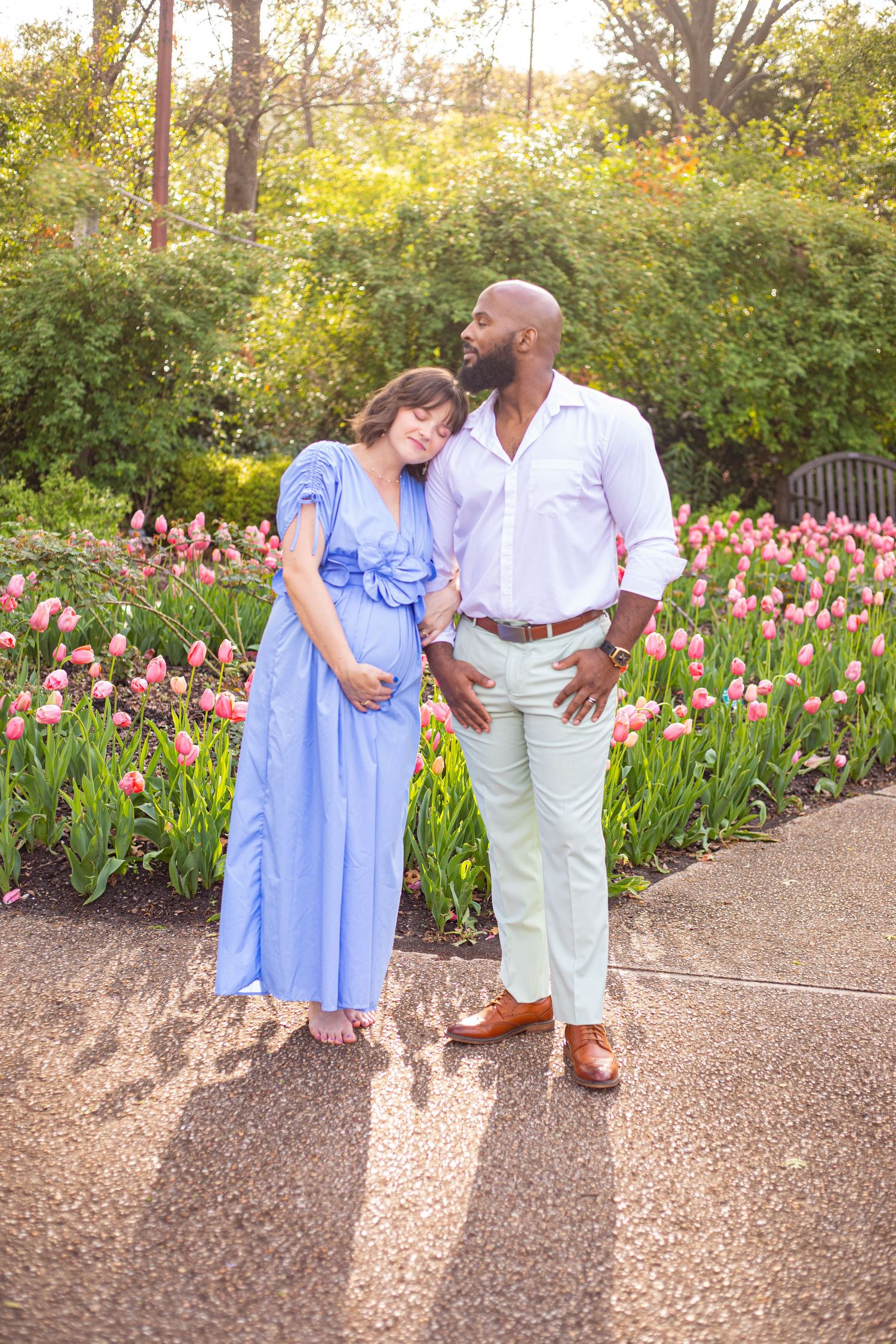 Couple shares intimate moment among pink tulips during outdoor maternity photoshoot.