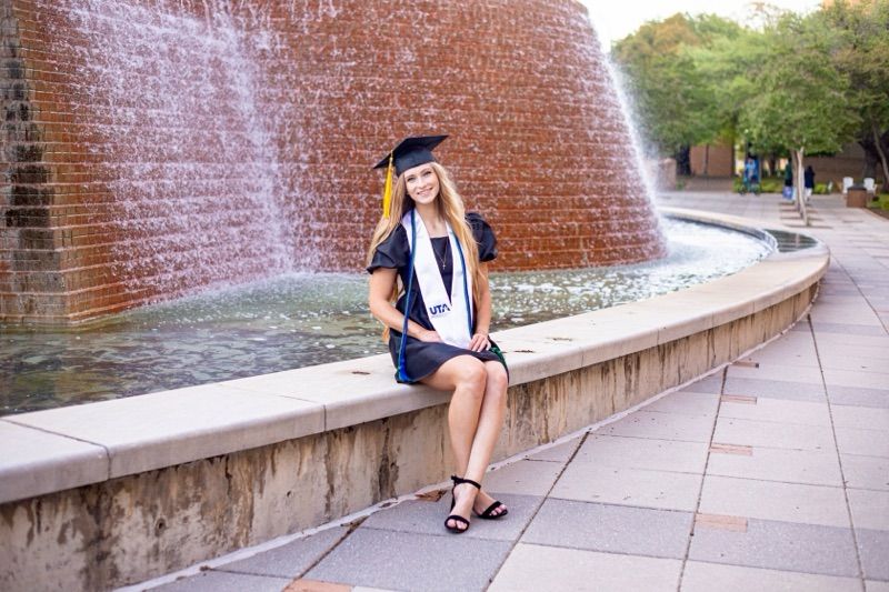 Graduate in cap and gown poses near a curved brick fountain wall on campus.