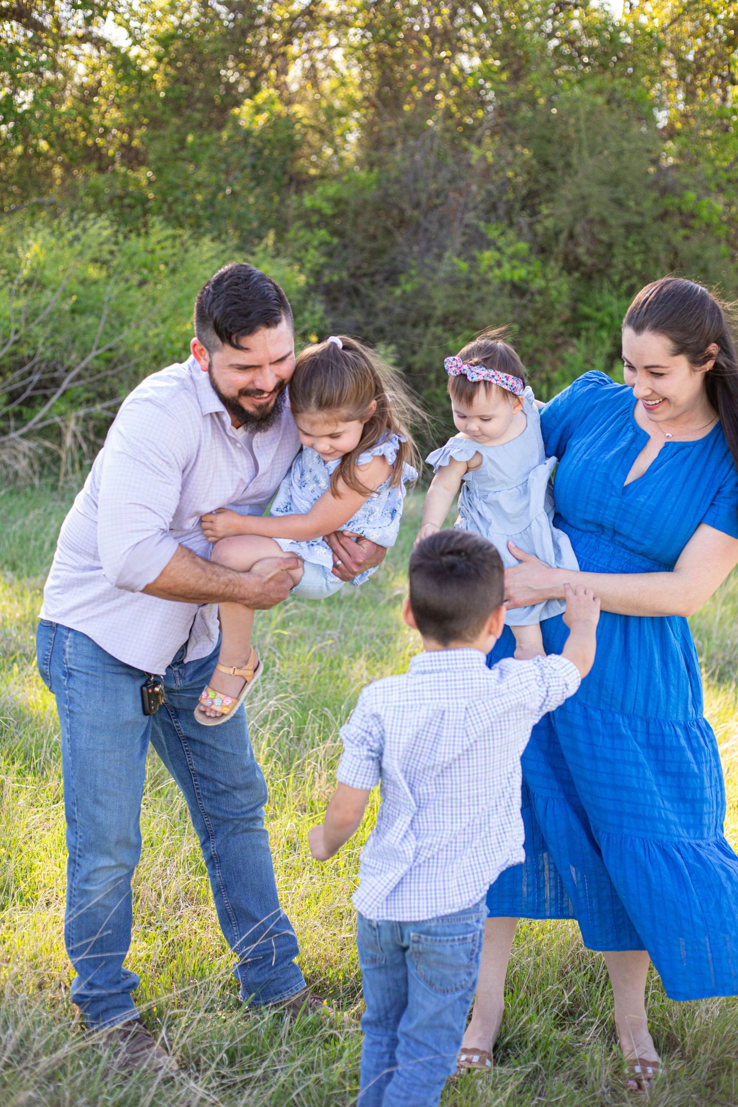 Happy family of five playing outdoors in green field, parents holding young children while toddler reaches up joyfully.