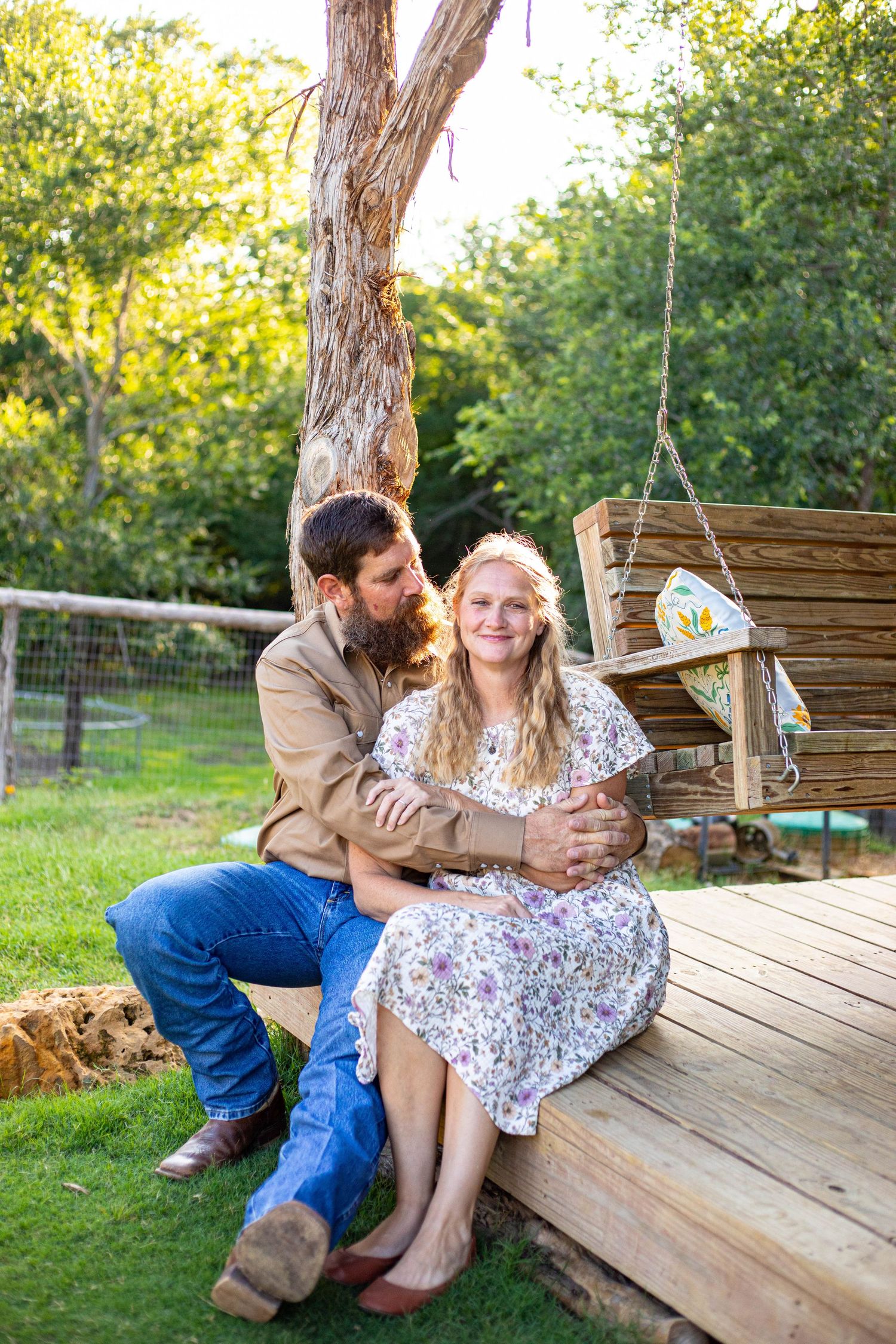 A couple sits together on a wooden deck near a tree swing, the man embracing the smiling woman in a floral dress.