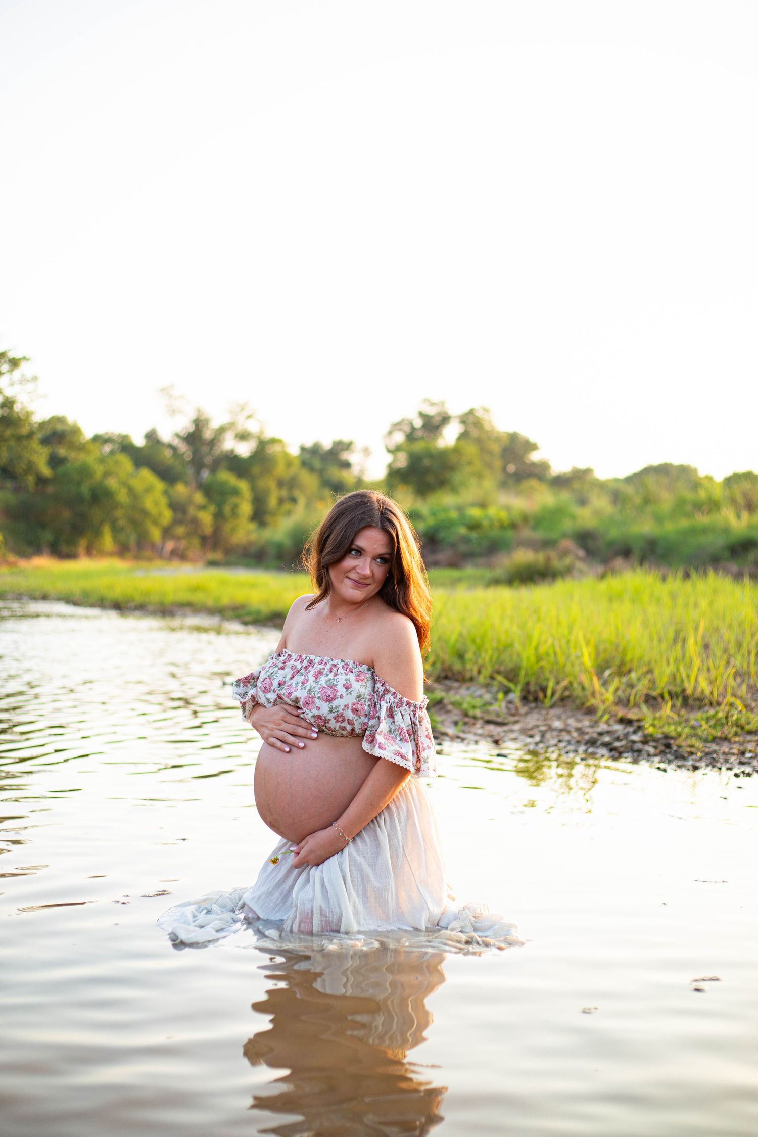 Pregnant woman in floral off-shoulder top stands in shallow water, cradling her baby bump during a golden hour maternity shoot.