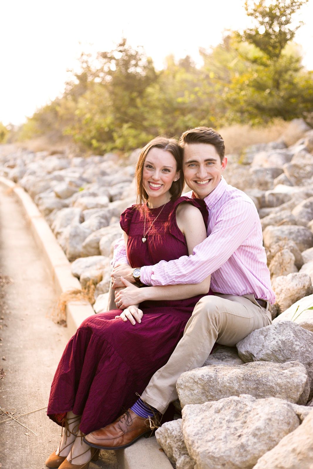 A couple sits together on rocky terrain at sunset near water.