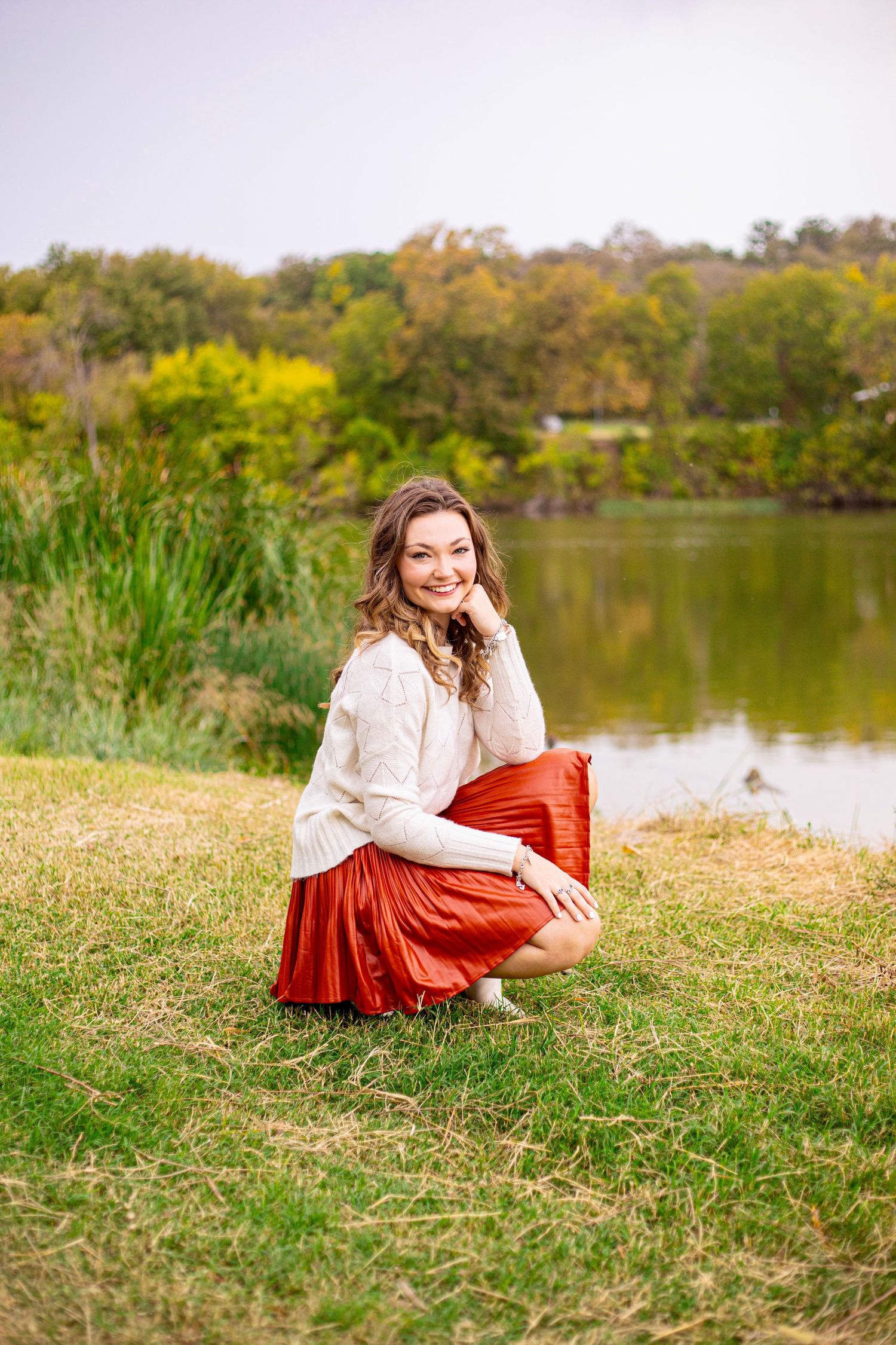 A person in a white sweater and red skirt sits by a serene lake surrounded by autumn foliage.