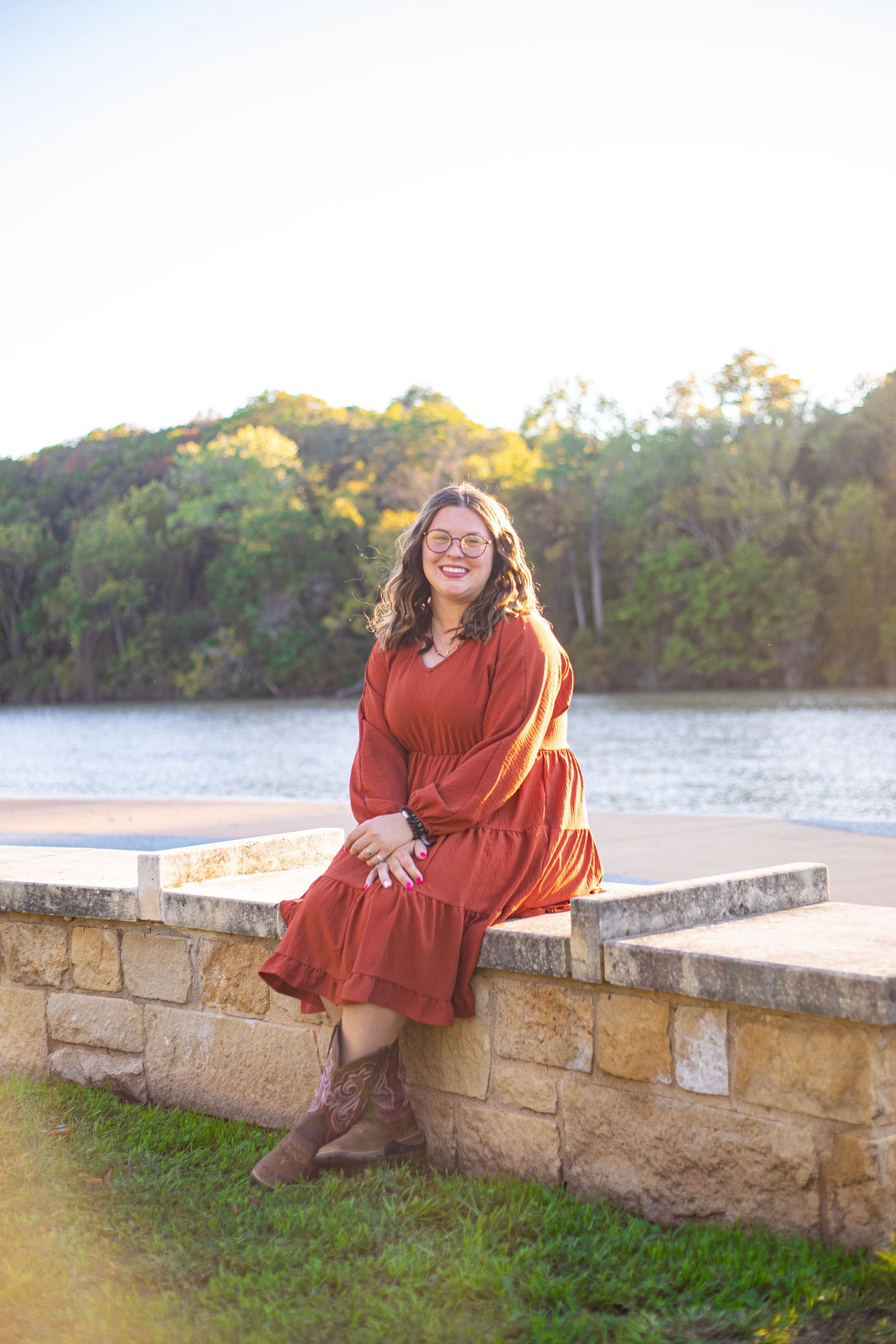 A person in a rust-colored dress sits on a stone wall overlooking a lake at sunset.