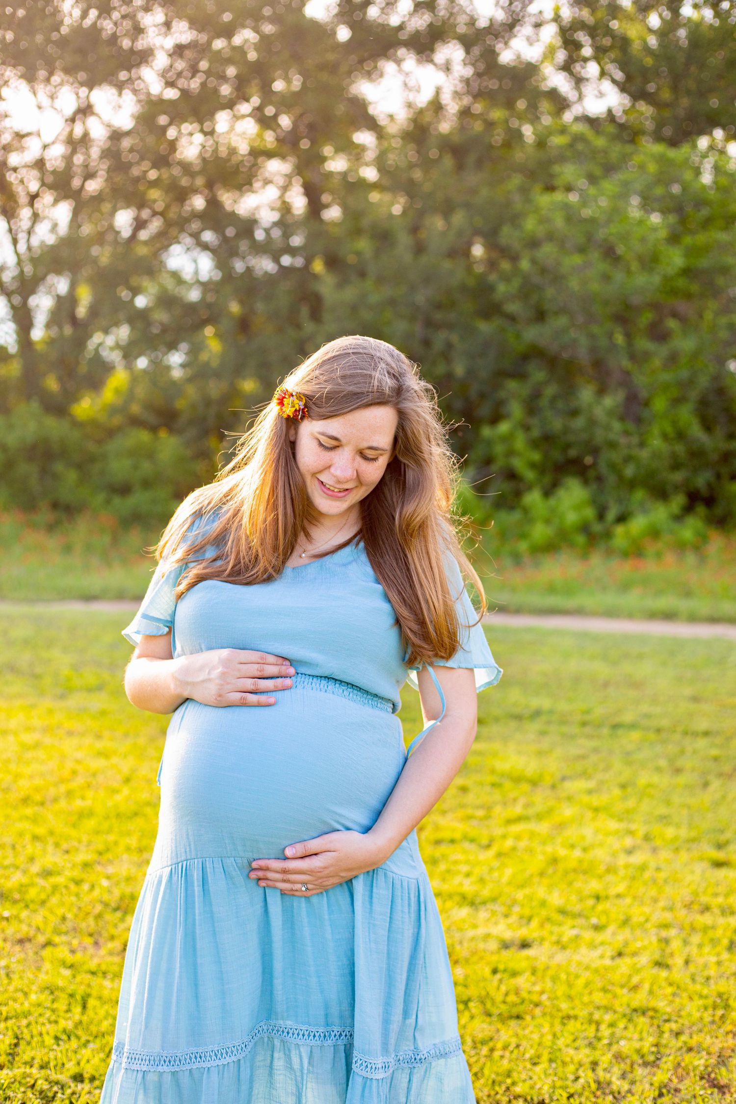 Pregnant woman in blue dress holding baby bump outdoors in sunny green park, smiling down with flower in hair.