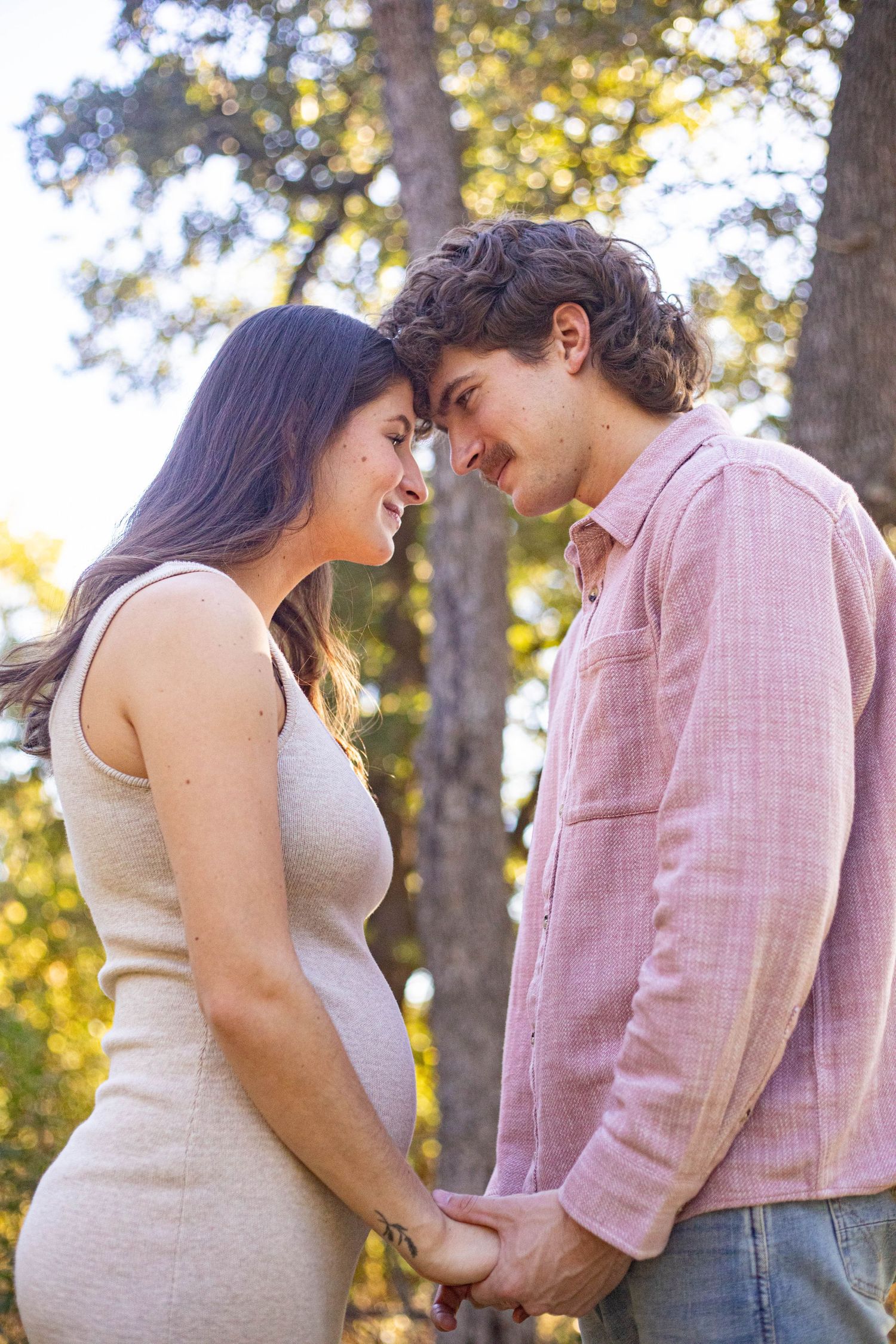 Pregnant couple holding hands and touching foreheads tenderly in a sunlit park setting.