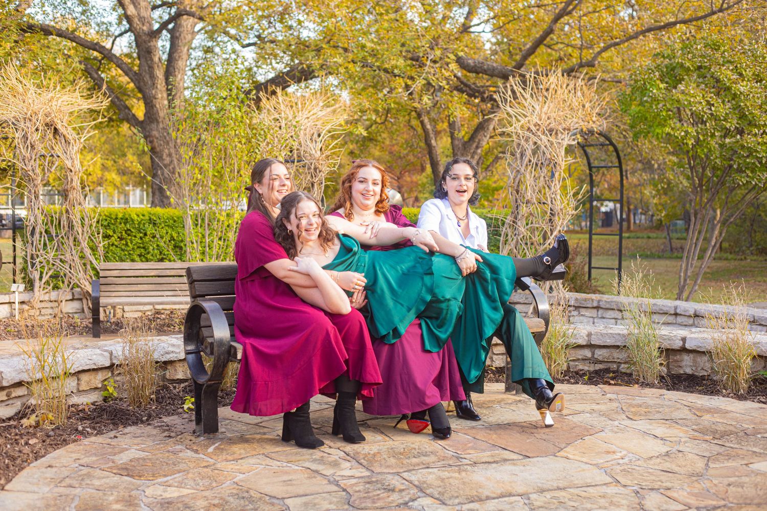 Four women in colorful Renaissance-style gowns pose together on a stone bench surrounded by autumn trees.