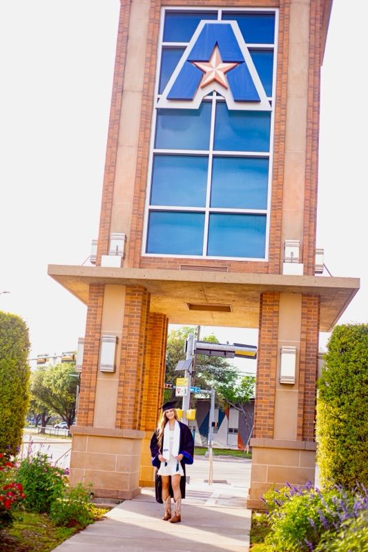 Graduate stands under architectural UTA logo entrance during graduation photo session.