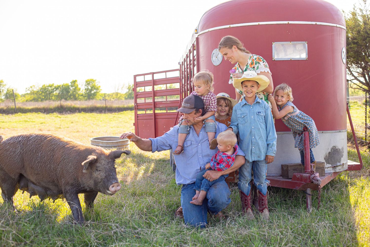 Several people interact with a large pig beside a red barn structure.