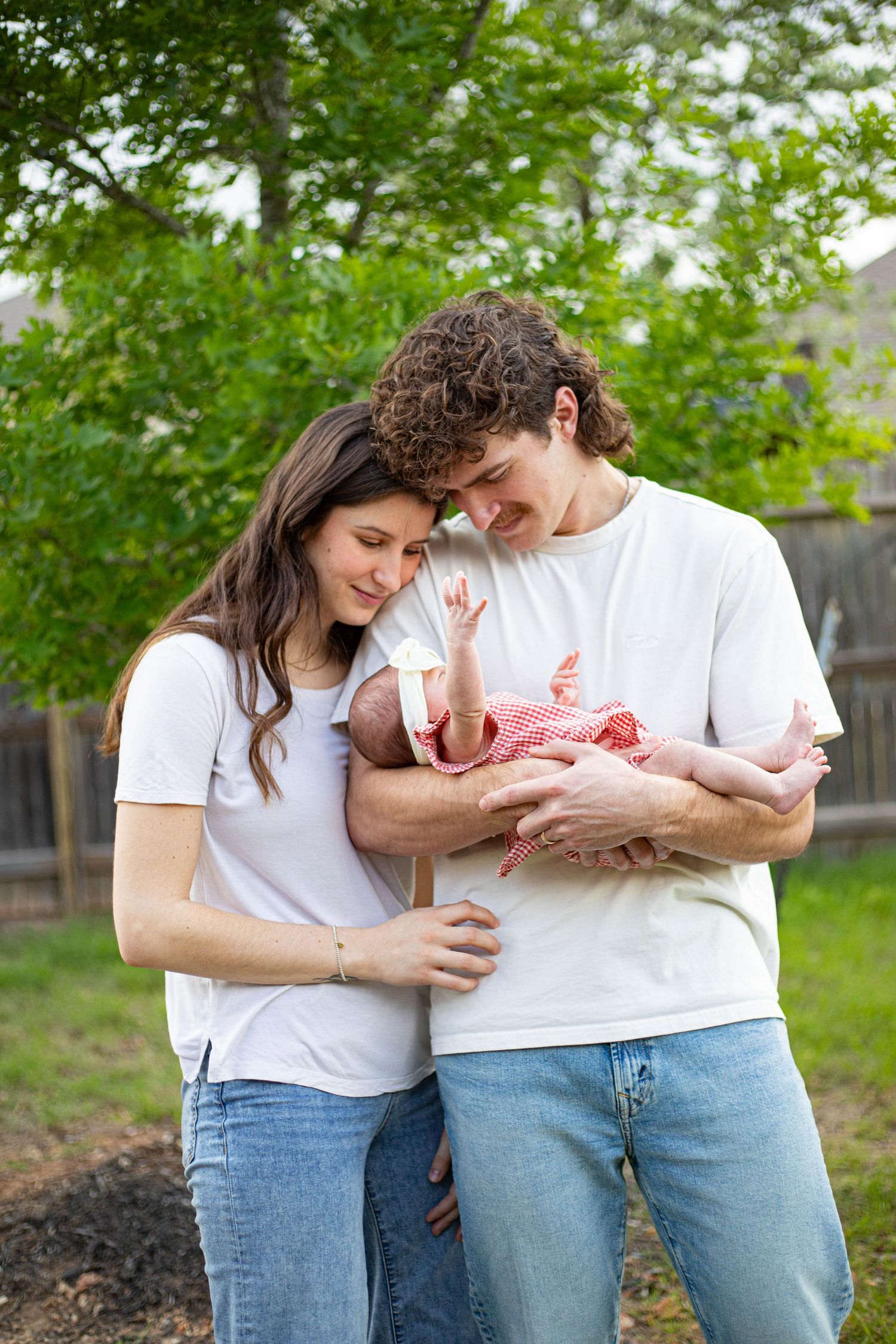 Young couple in white tees and jeans lovingly holding their newborn baby outdoors in a green backyard.