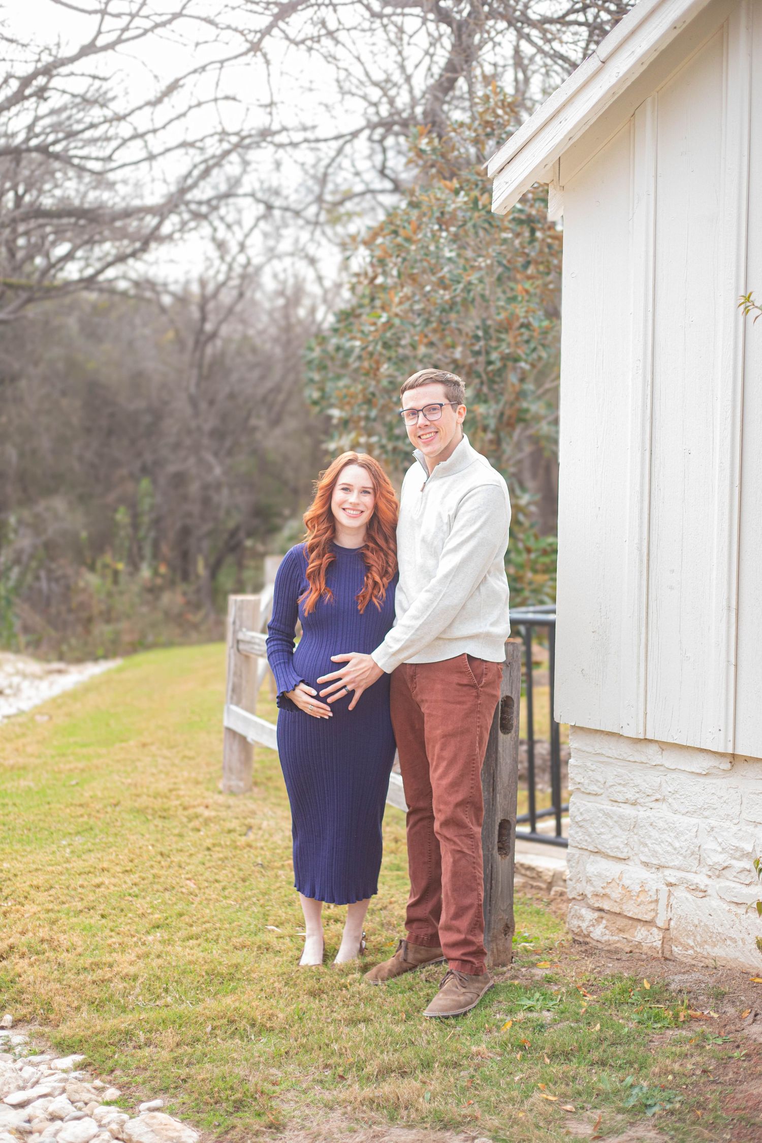 Pregnant couple posing outdoors near a white building, woman in blue dress, man in white shirt and rust pants.