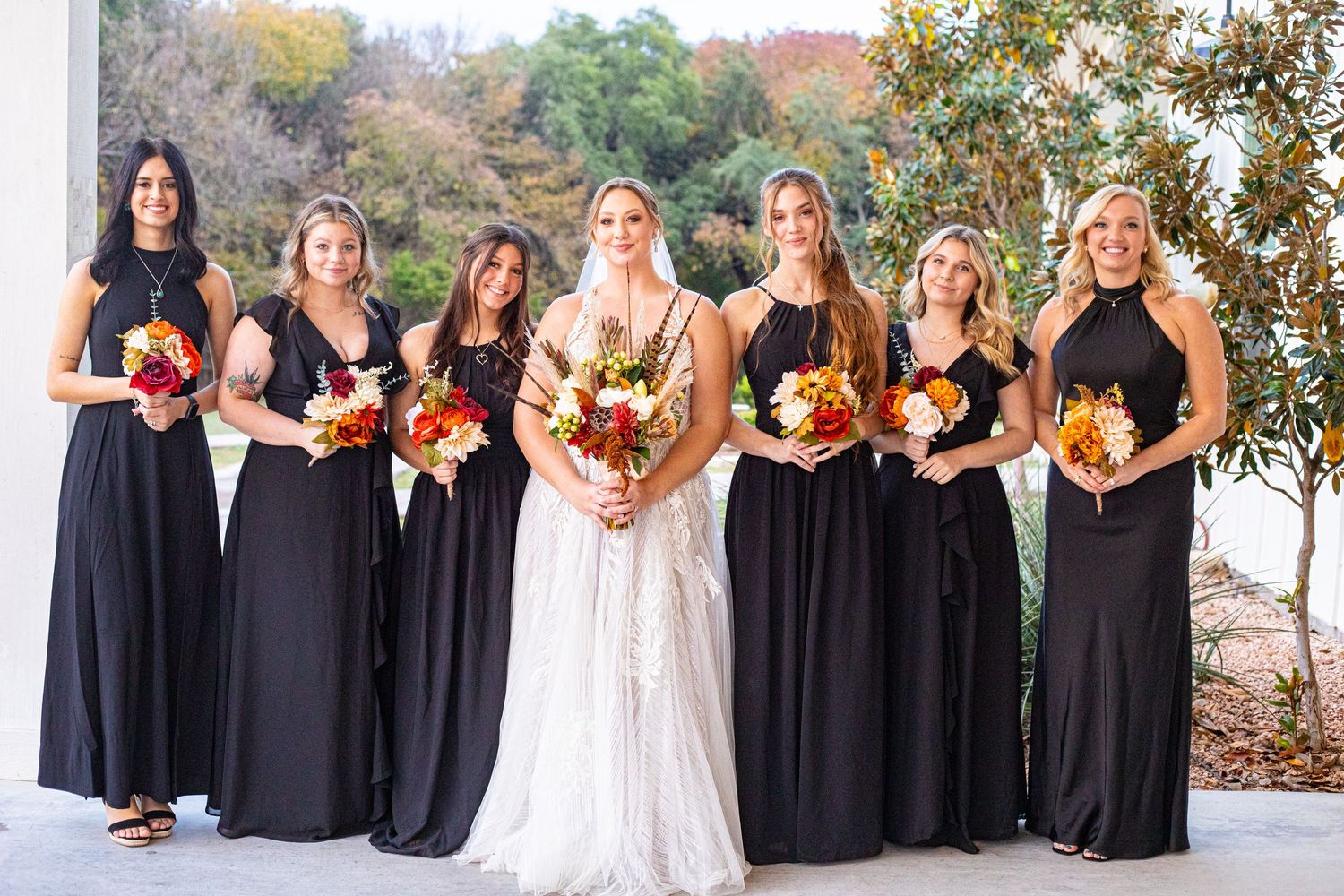 A bridal party holding orange and red bouquets poses in black dresses against an autumn backdrop.