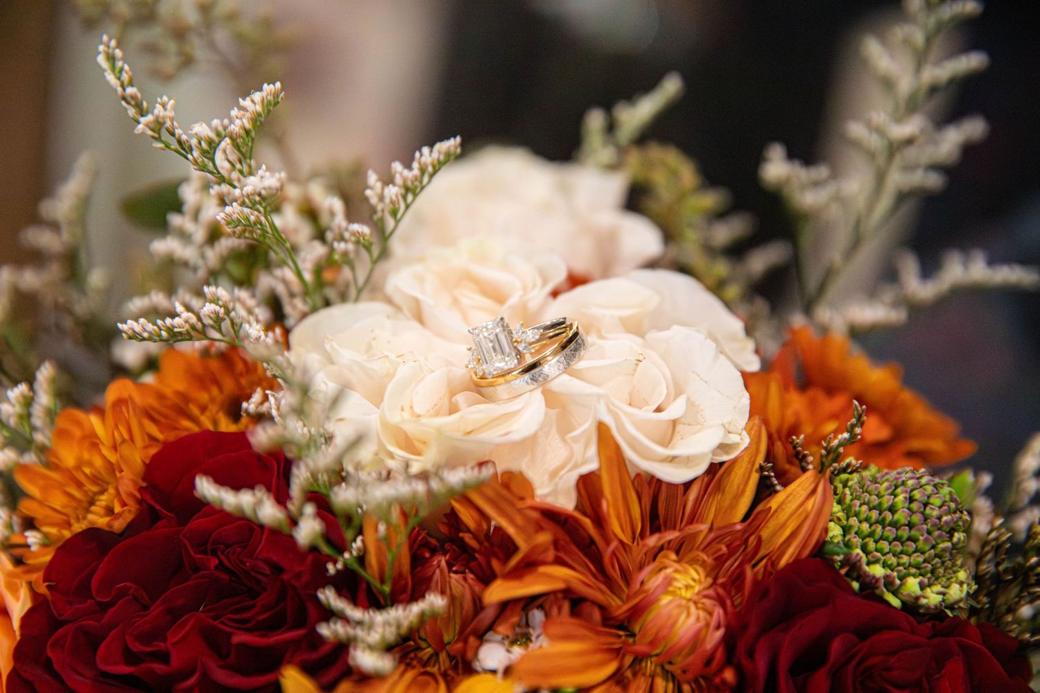Fall wedding bouquet with ivory roses, orange dahlias, and burgundy flowers, adorned with sparkling wedding rings.