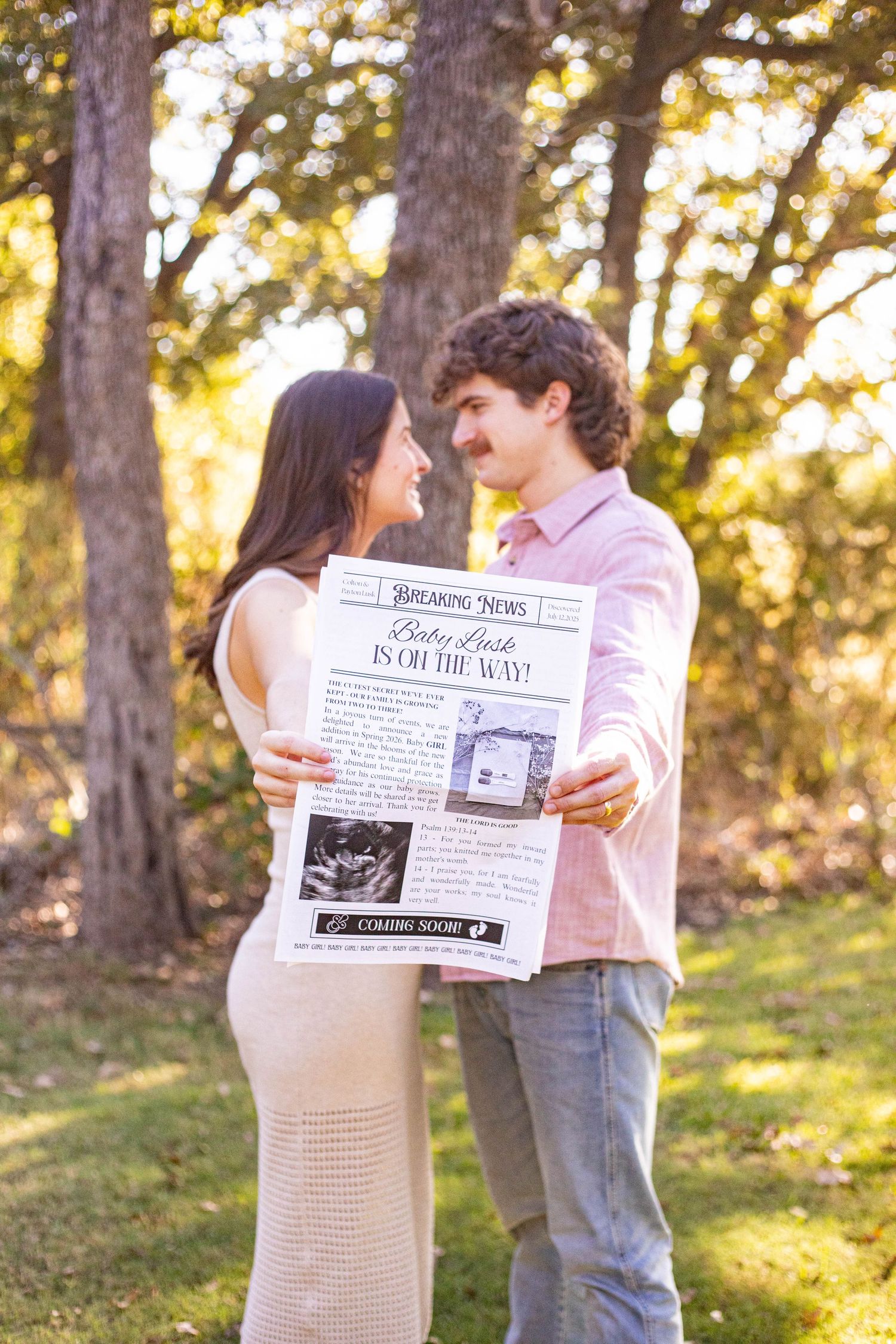 Couple holding a pregnancy announcement newspaper in a sunny park, smiling at each other, expecting baby news.