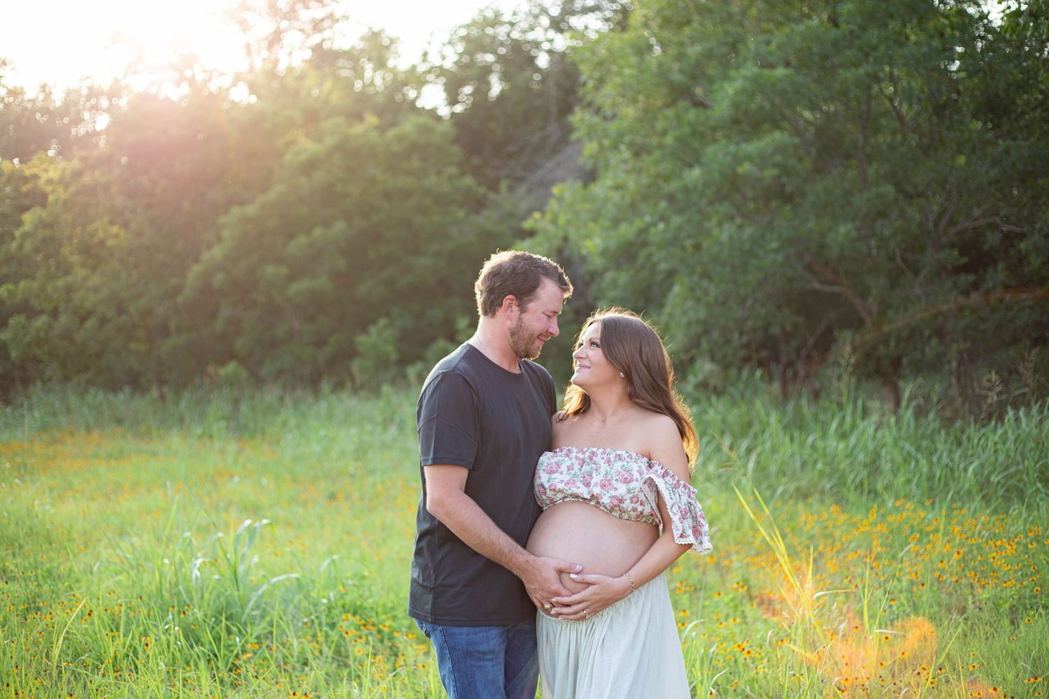 Couple sharing a tender moment outdoors at golden hour, woman in floral dress cradling pregnant belly in lush green field.