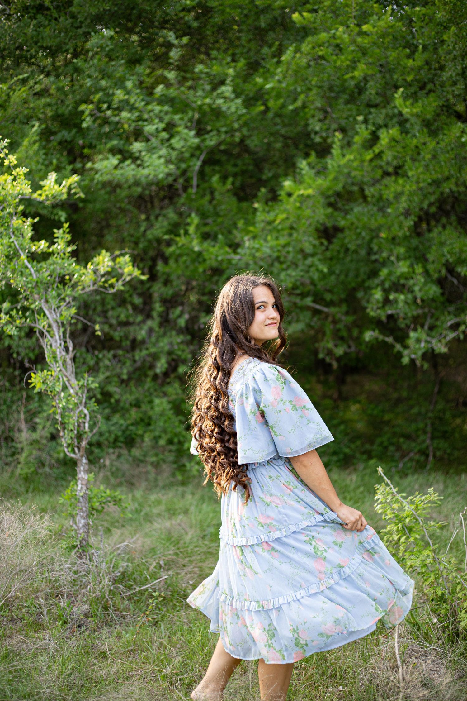Young woman in a light blue floral dress twirling in a lush green outdoor setting with long curly hair.