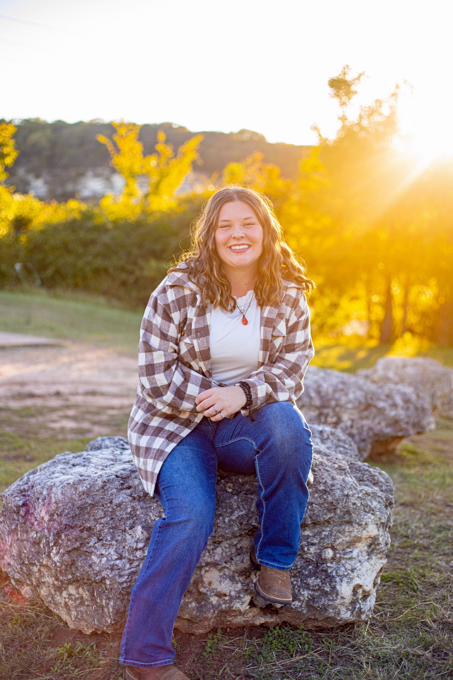 A person in plaid and denim sits on rocky terrain during golden hour sunset.