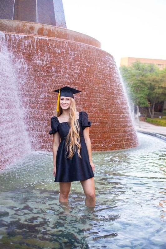 Graduate poses playfully in the campus fountain water wearing academic regalia.