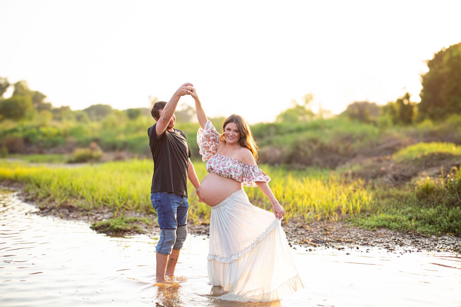Couple dancing in shallow water during golden hour, pregnant woman in white flowy skirt and floral top.