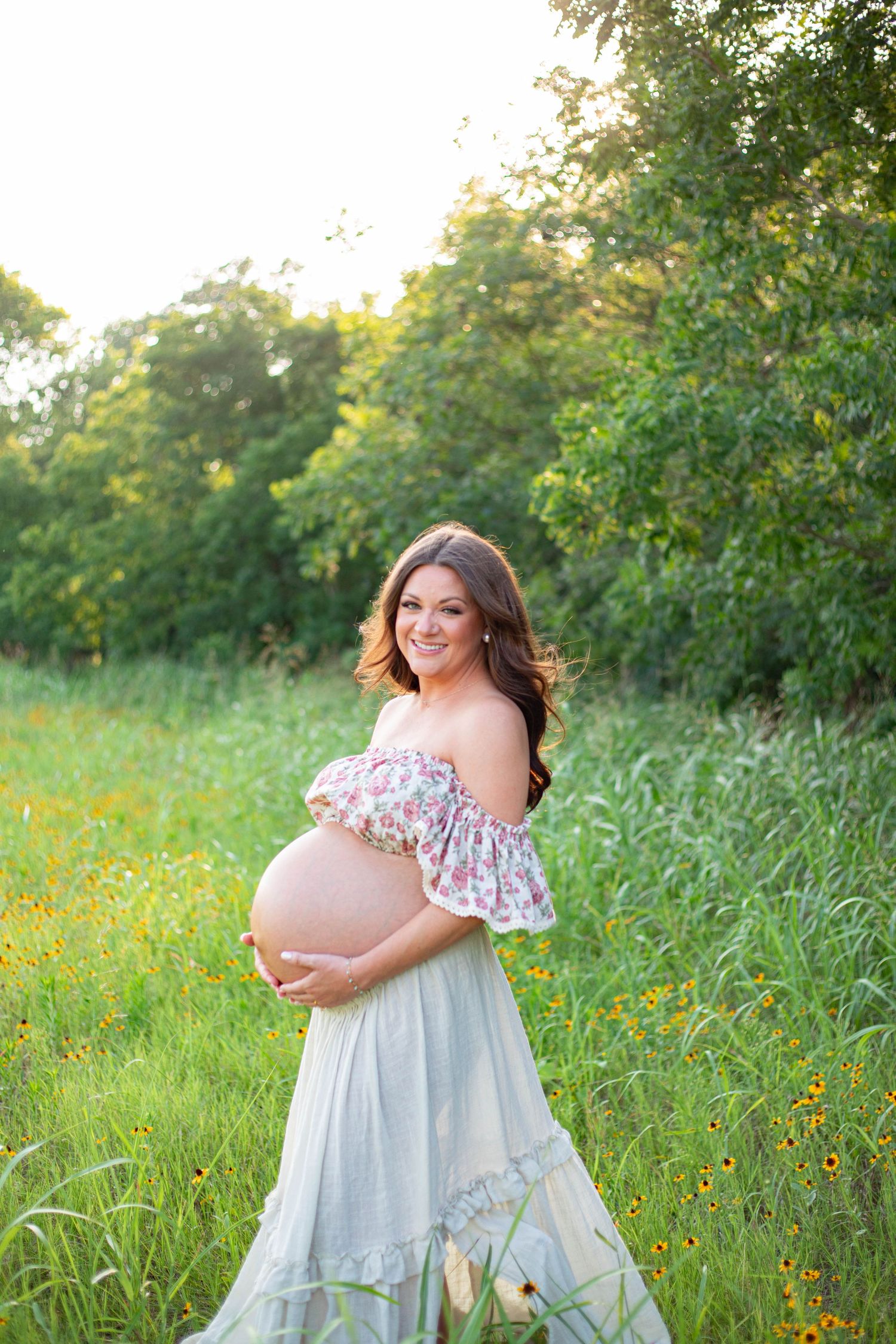 Smiling pregnant woman in floral off-shoulder top and mint skirt cradling baby bump in sunlit wildflower meadow.