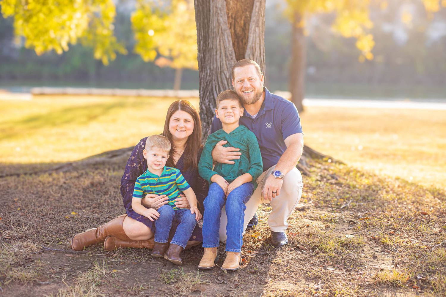 Family of four poses together under a tree in a sunny autumn park, parents and two young boys smiling warmly.