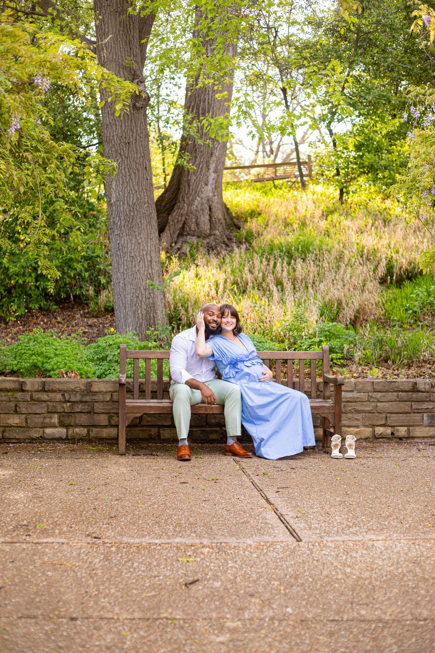 A couple sits together on a stone bench in a lush garden setting during a maternity photoshoot.