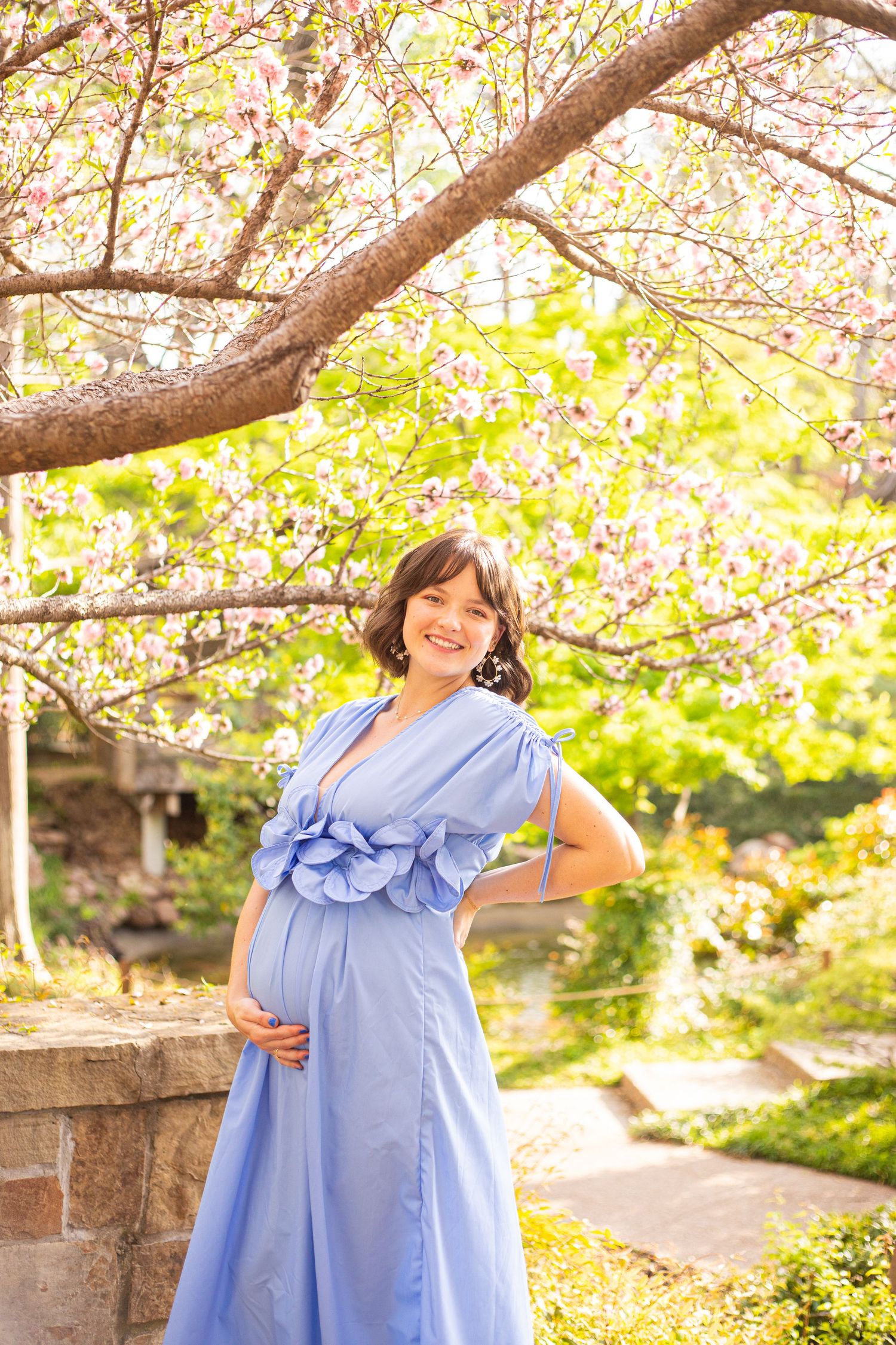 A woman in a flowing blue dress poses under blooming cherry blossoms in a garden setting.