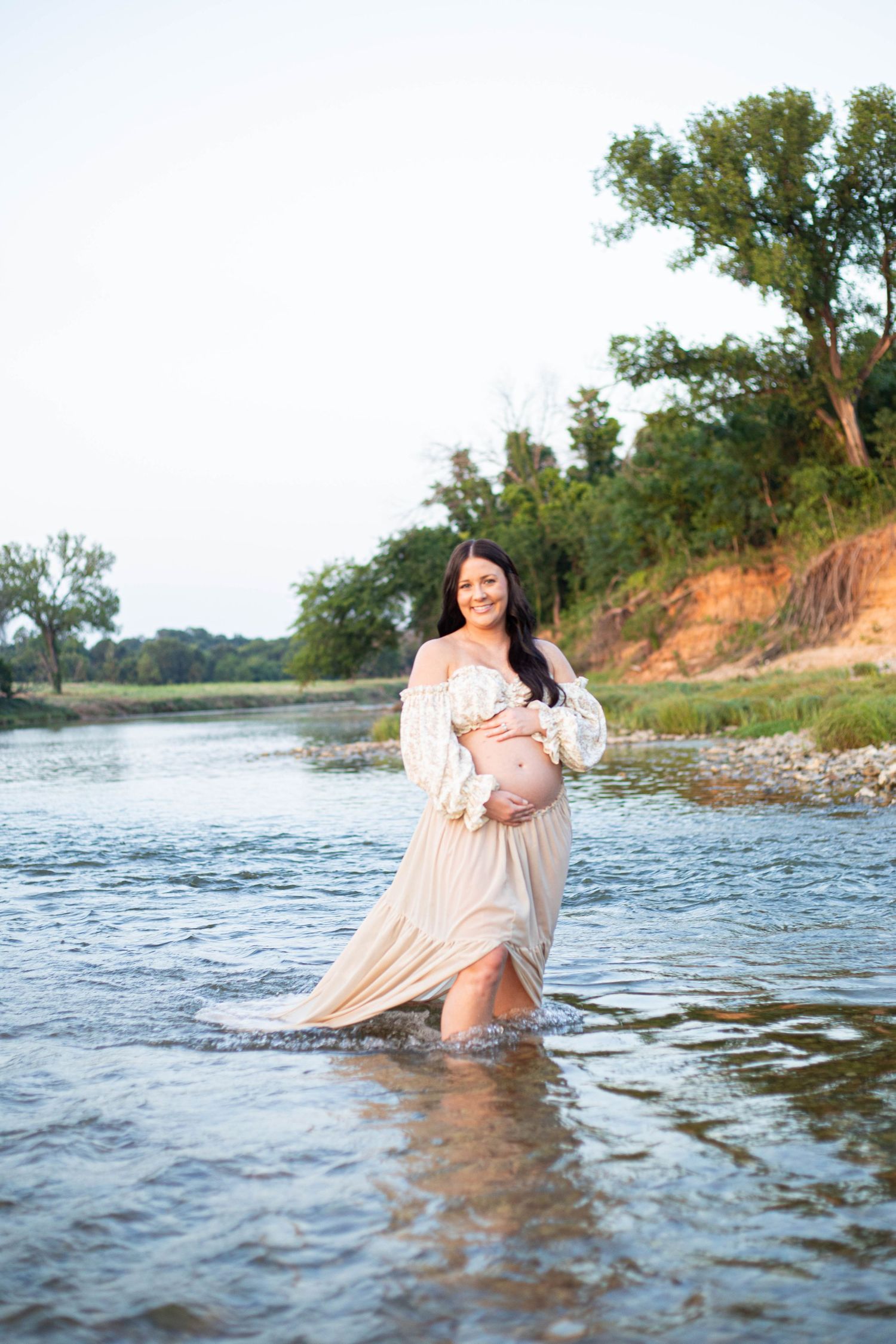 Pregnant woman in flowing beige dress standing in a shallow river, smiling and cradling her baby bump outdoors.